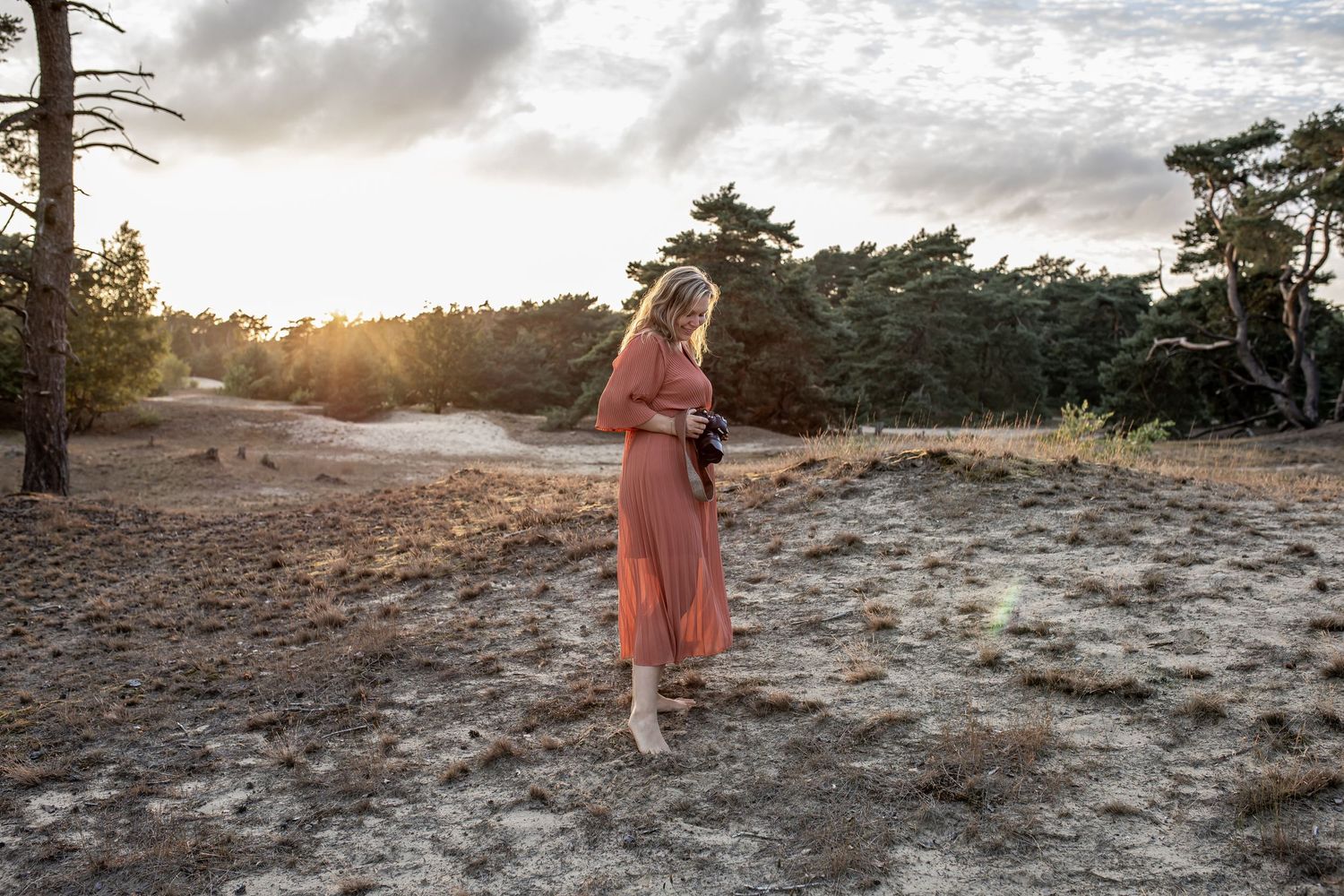 Woman in pink dress standing on sandy heathland at golden sunset with pine trees behind.