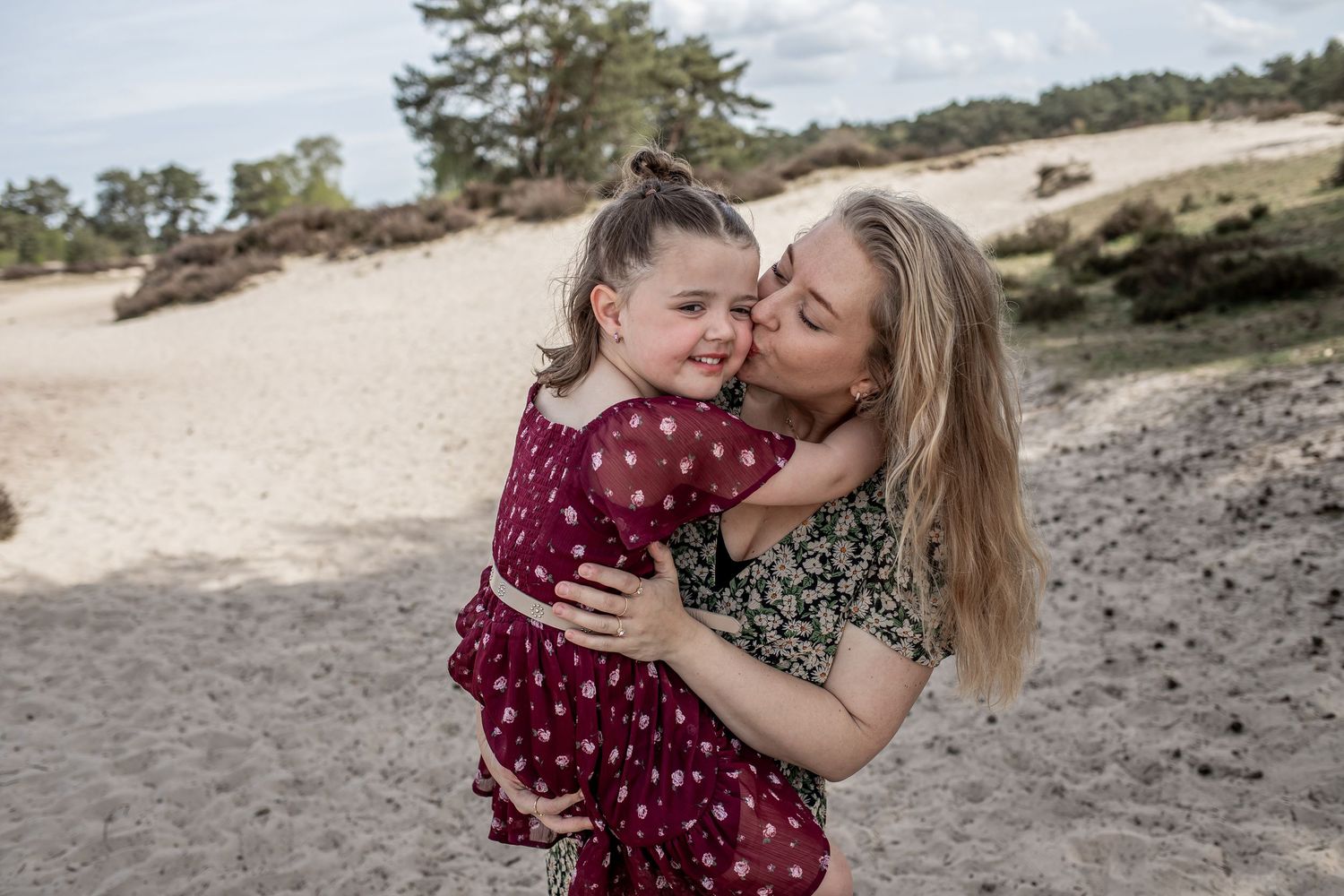A mother lovingly kisses her smiling young daughter while holding her on a sandy beach surrounded by trees.