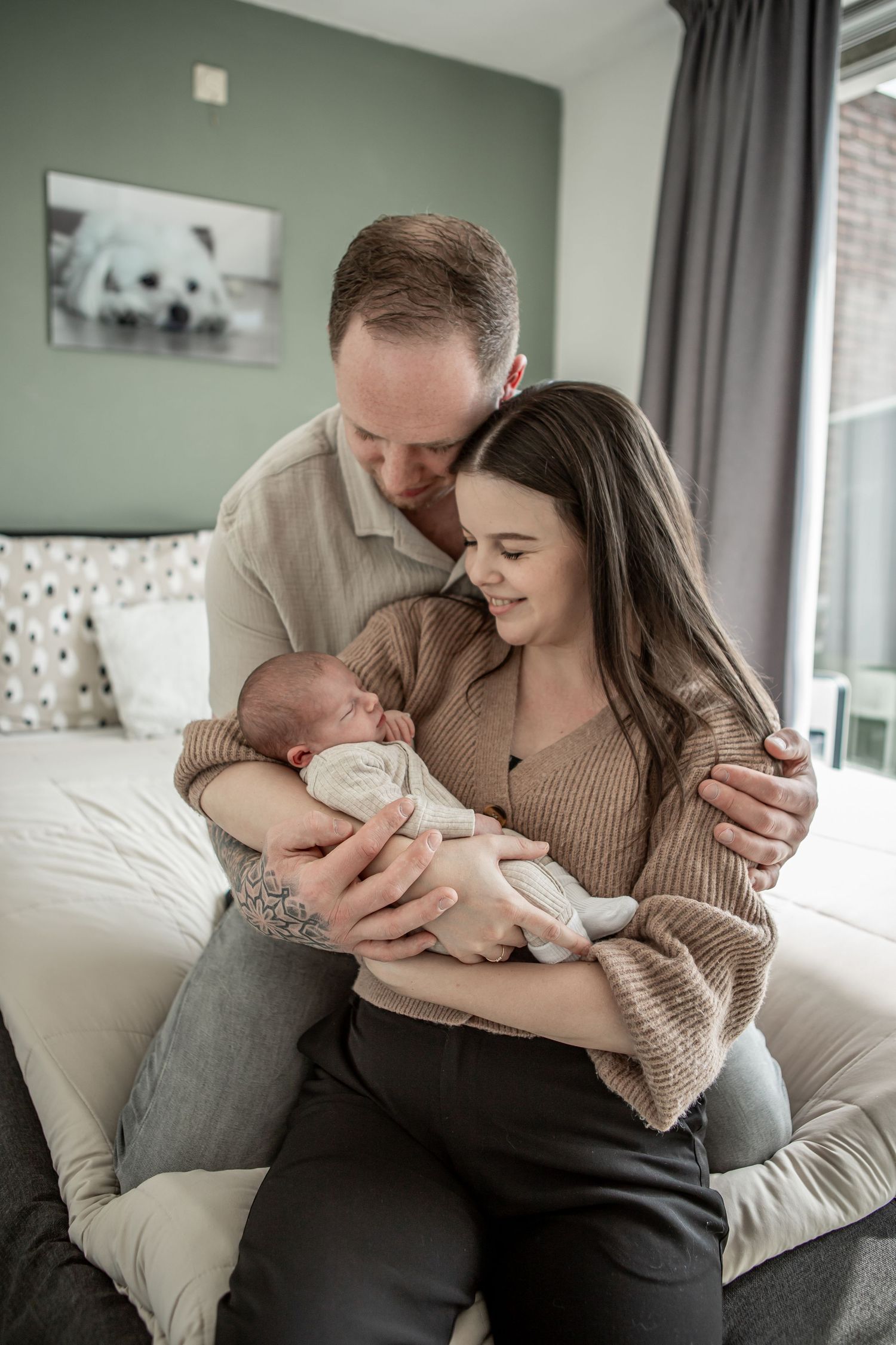 New parents sitting on a bed lovingly gazing at their newborn baby in a cozy bedroom.