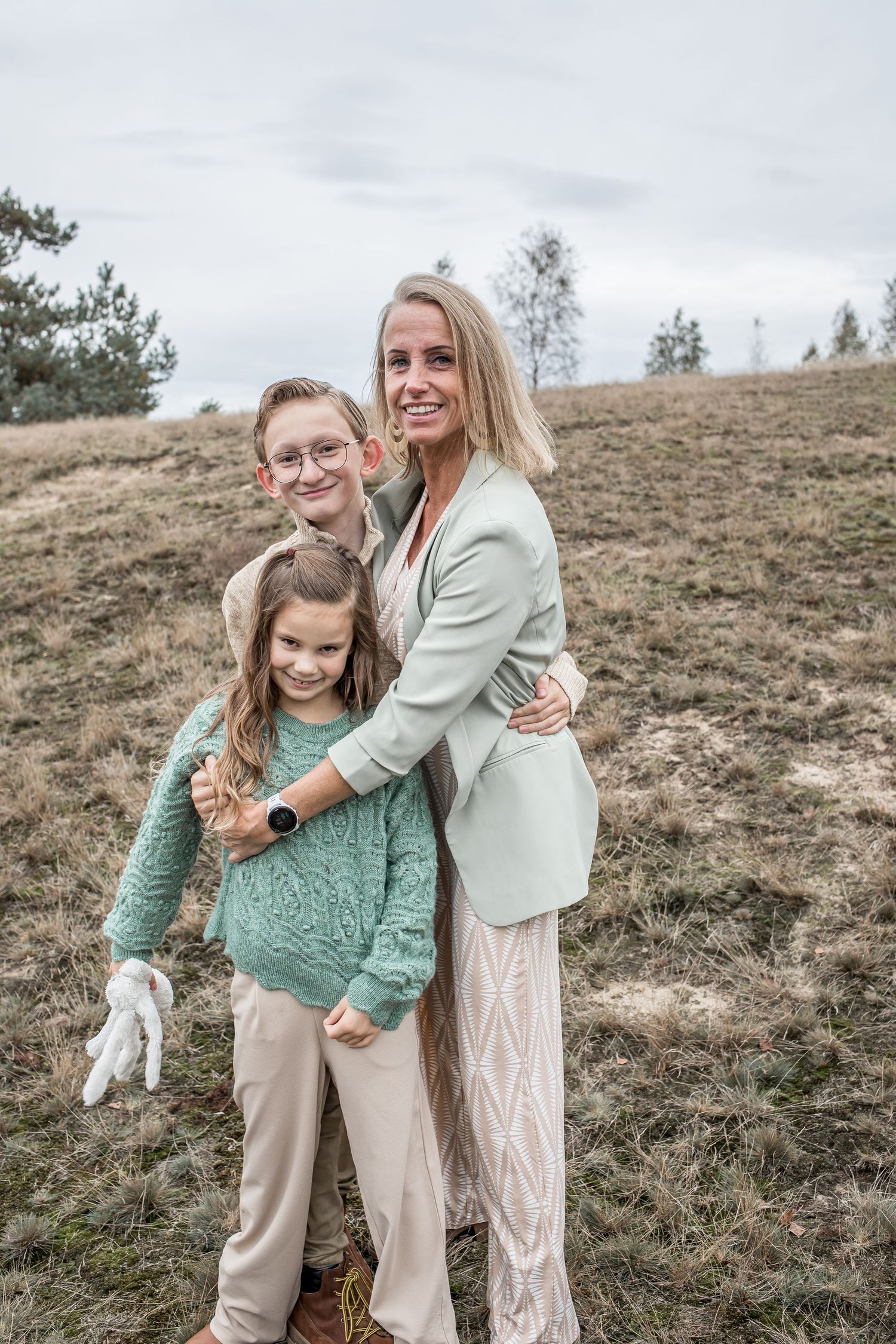 A blonde woman stands with two children on a grassy hillside on a cloudy day.