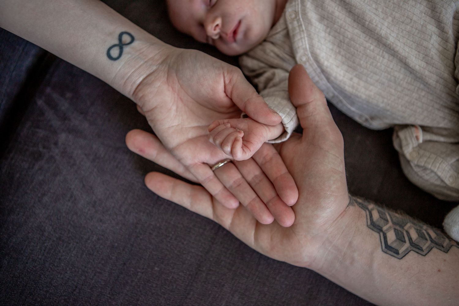 Two adult hands cradling a sleeping newborn baby's tiny hand, with infinity and geometric tattoos visible.