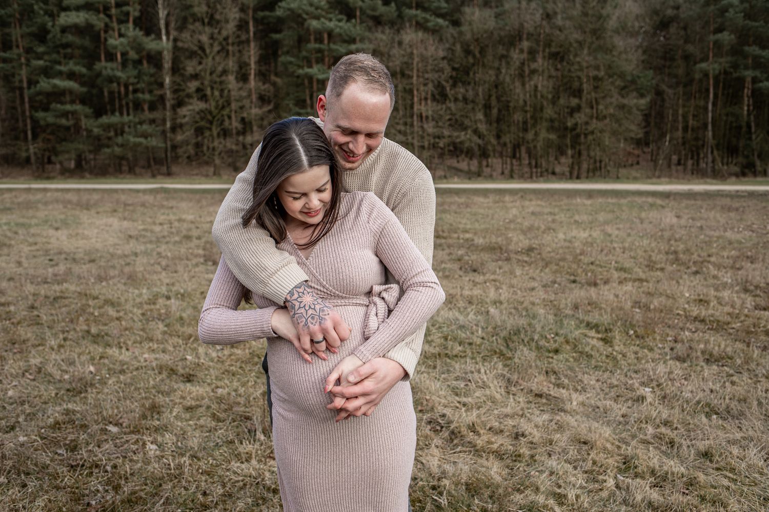 Pregnant couple embracing outdoors in a field, woman in fitted dress with partner holding her belly.