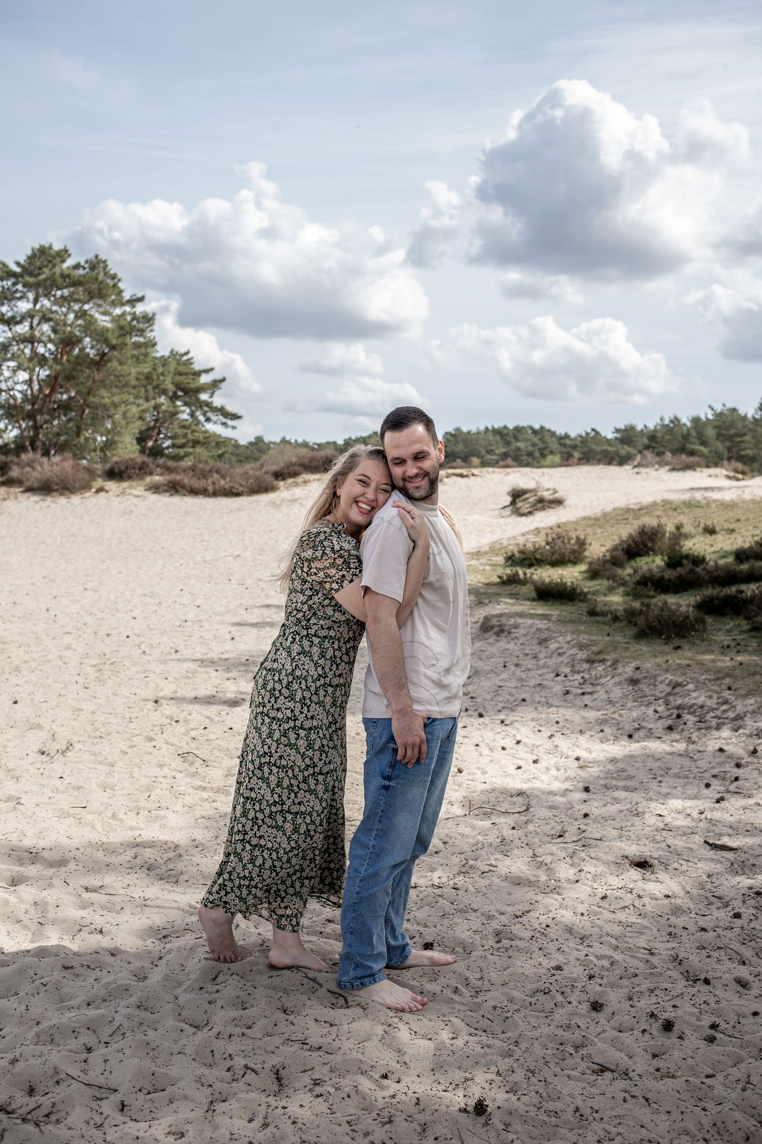 A couple embracing on sandy dunes, woman in floral dress hugging man in jeans, trees and cloudy sky behind them.