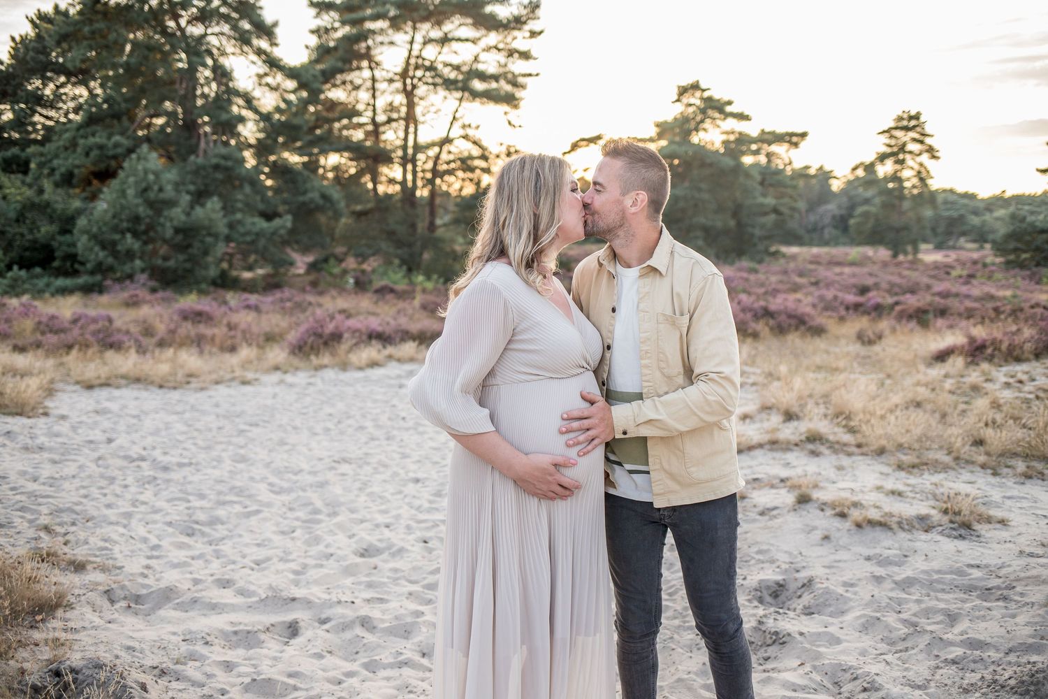 Pregnant couple kissing on sandy heath landscape at golden hour, woman in flowing beige maternity dress.