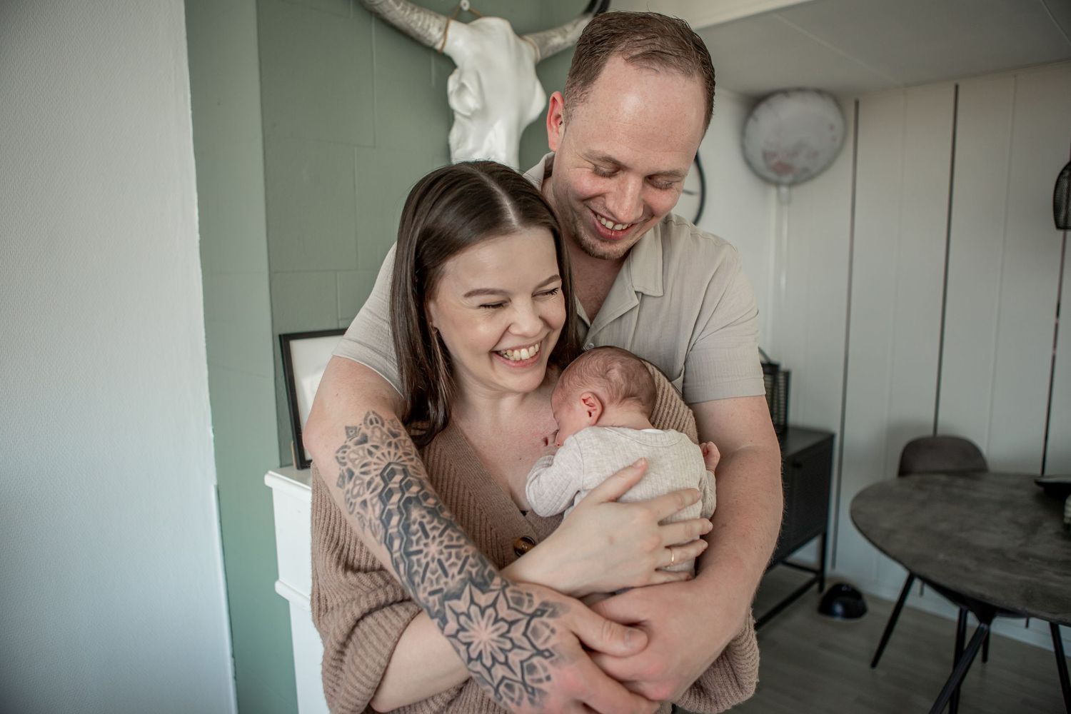 Joyful new parents laughing and cuddling their newborn baby in a green-walled room.
