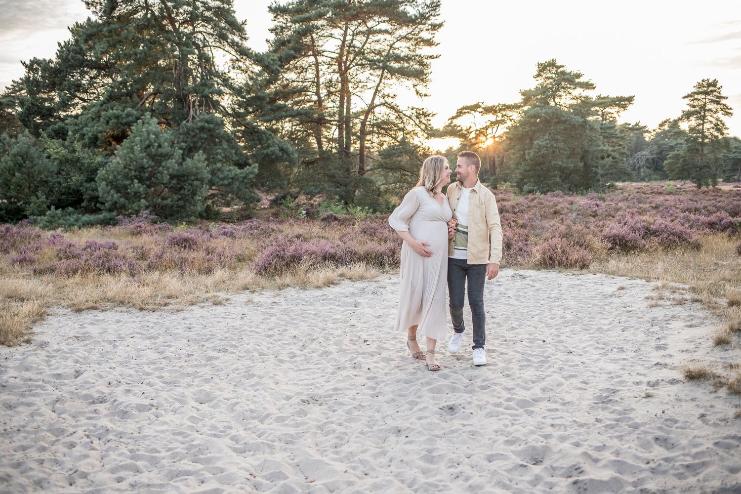 Couple strolling together on a sandy path surrounded by purple heather at golden sunset.