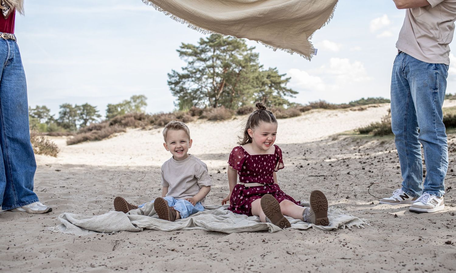 Two young children sit on a blanket on a sandy beach dune, smiling, while adults stand nearby holding a canopy.