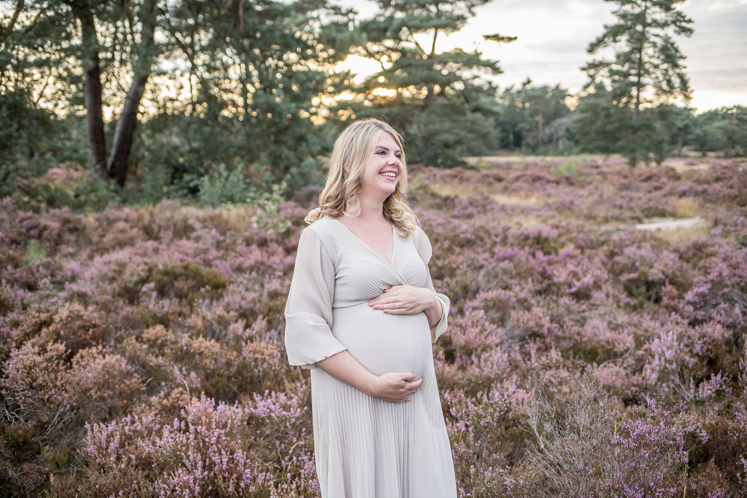 Smiling pregnant woman in a flowing grey maternity dress standing in a blooming heather field.