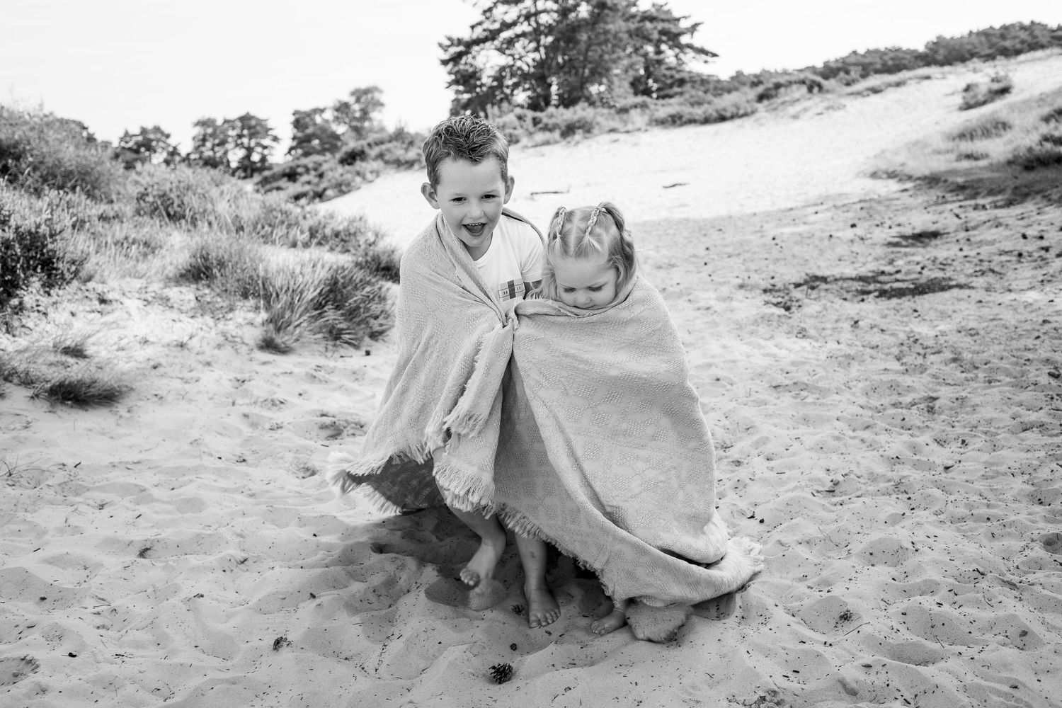 Two children laugh together wrapped in a blanket on a sandy beach in a black and white photo.