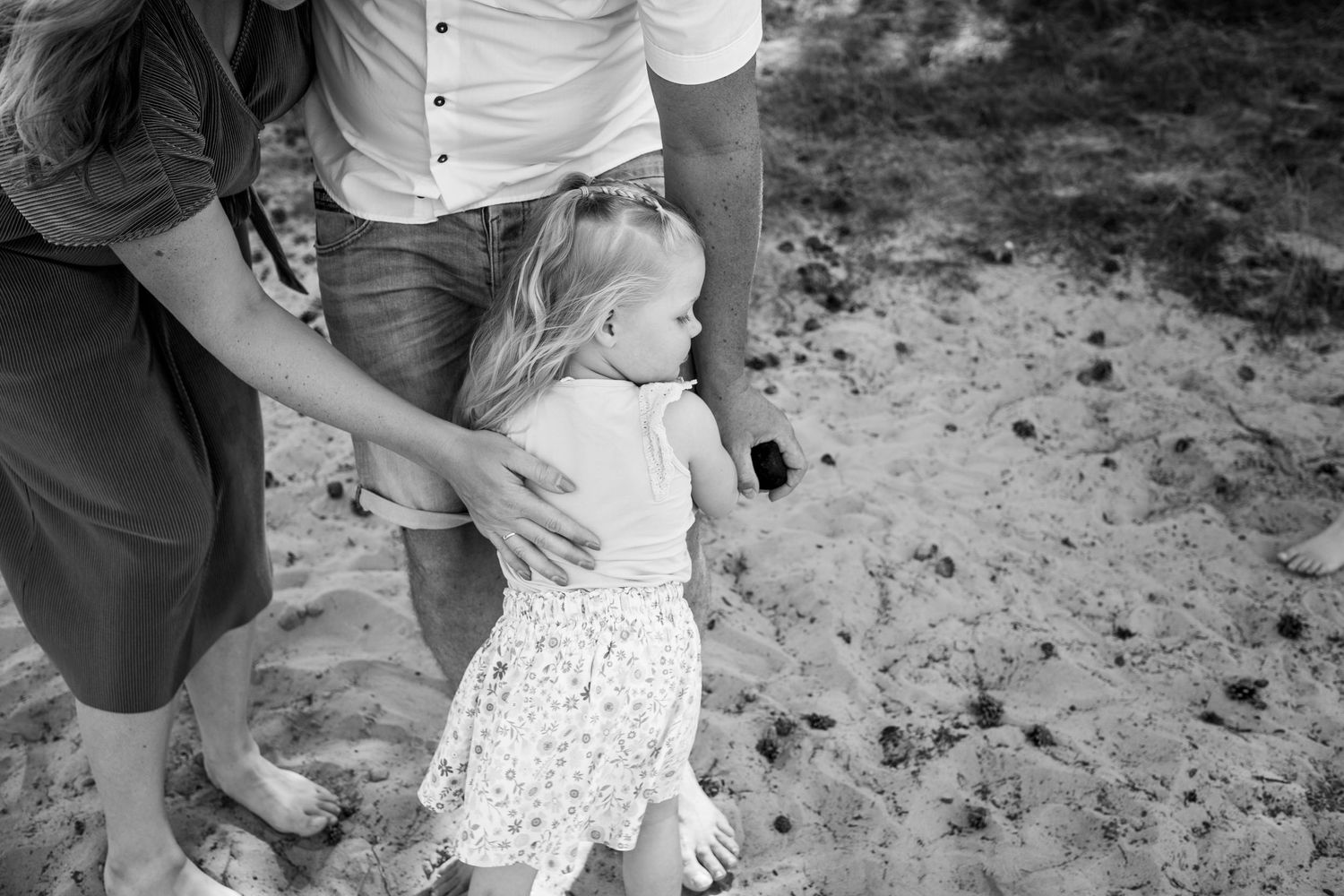 Black and white photo of parents helping their toddler daughter walk on sandy beach.