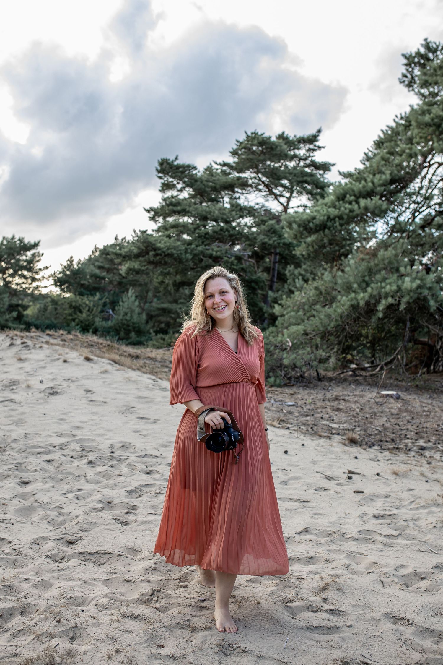 Smiling blonde woman in rust pleated dress holding camera on sandy dunes.