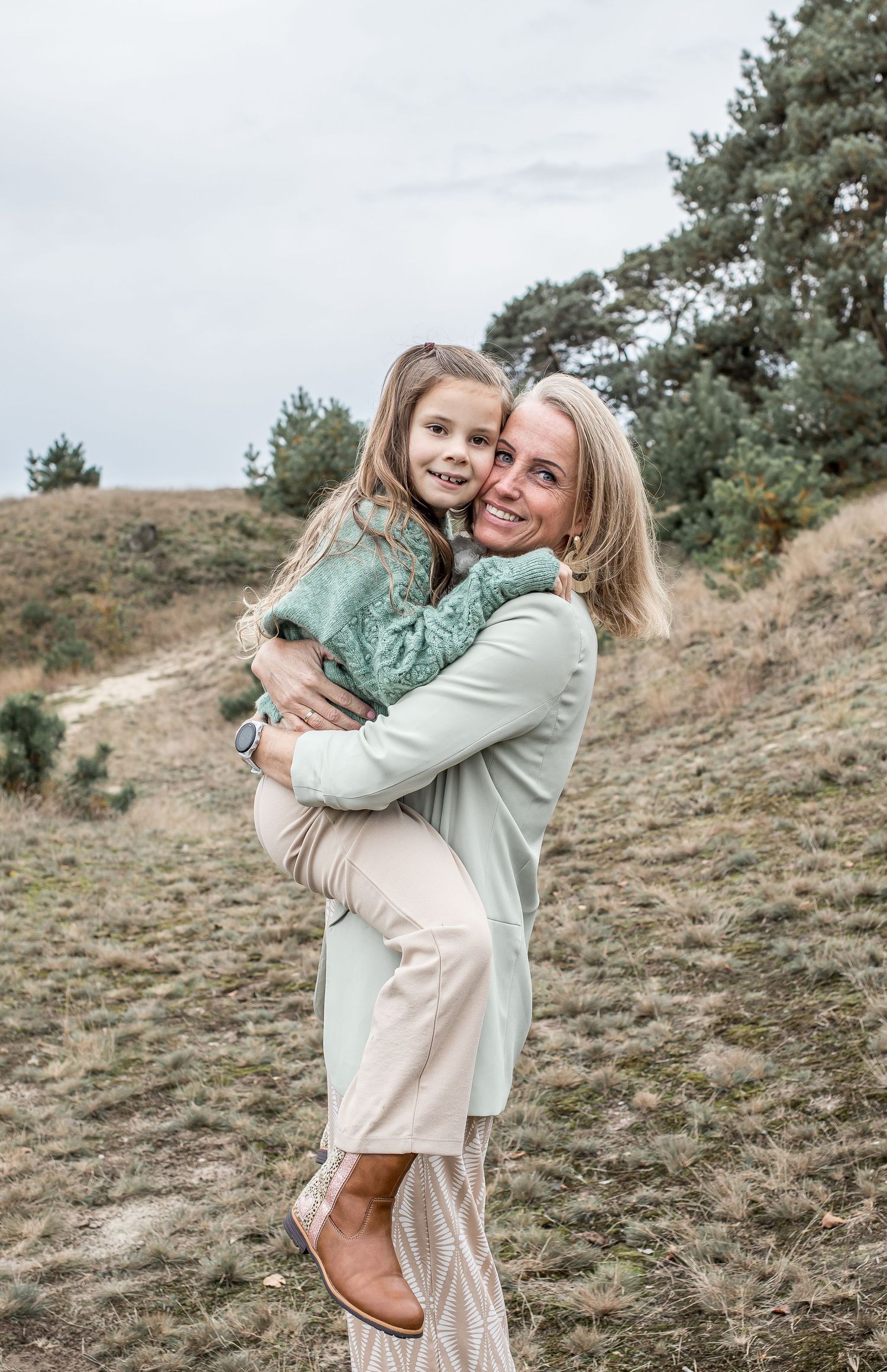 A smiling blonde woman holds a young girl in a green sweater on a grassy hillside.