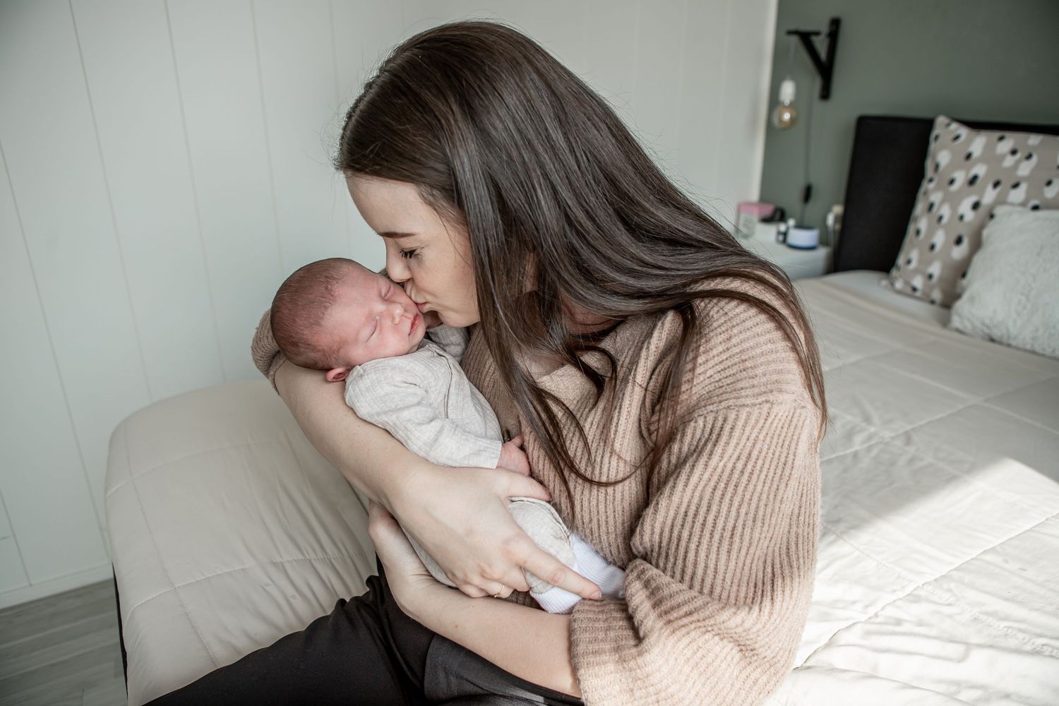 Dark-haired mother gently touching noses with her swaddled newborn baby in a cozy bedroom.
