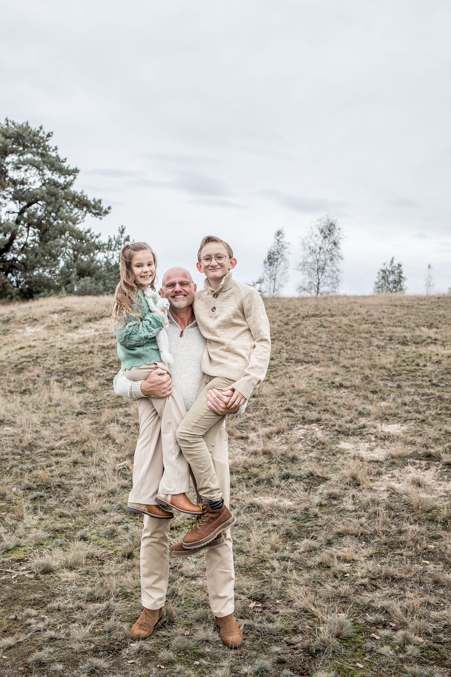 A man holds two children in his arms while standing in an open grassy field outdoors.