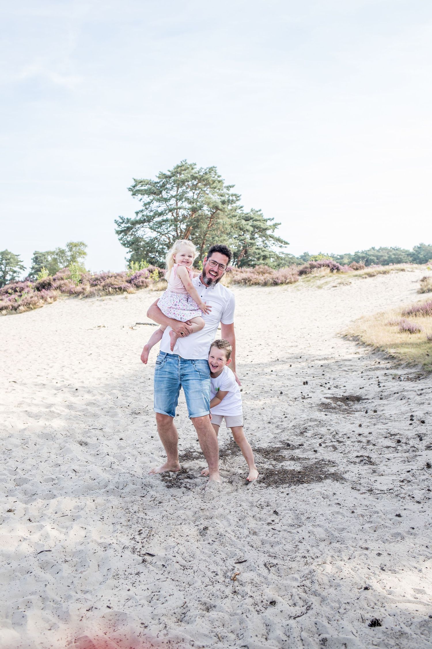 A laughing father holds his young daughter while his son playfully leans against him on a sunny sandy dune.