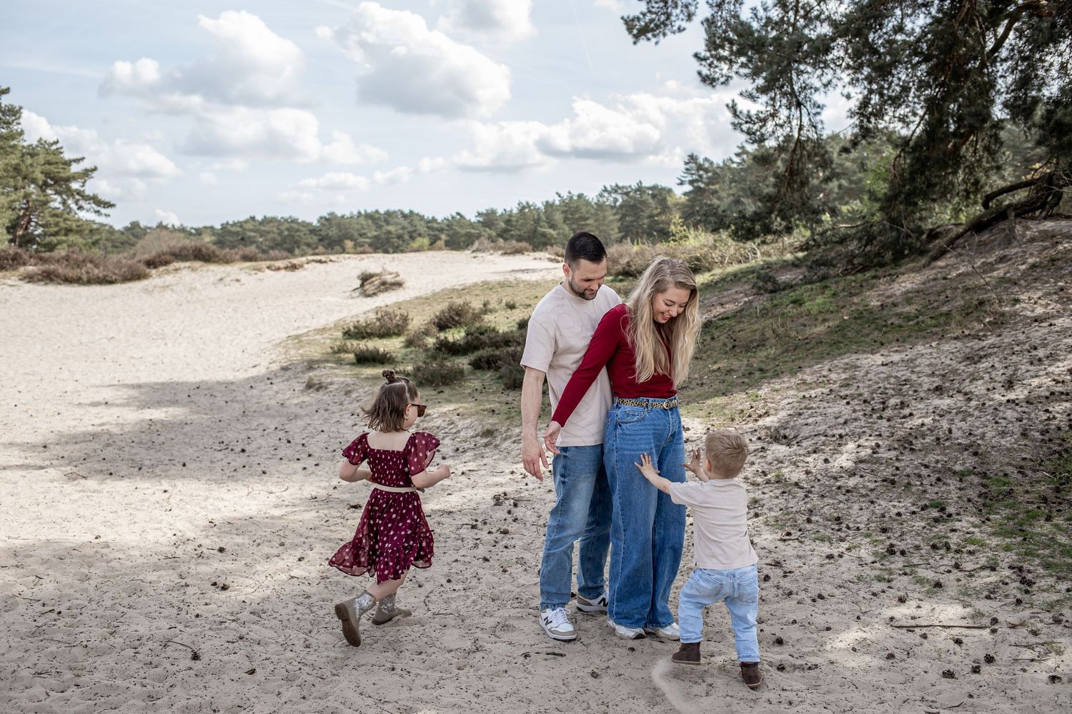 Family of four walking on sandy path with pine trees, parents holding hands with toddlers outdoors.