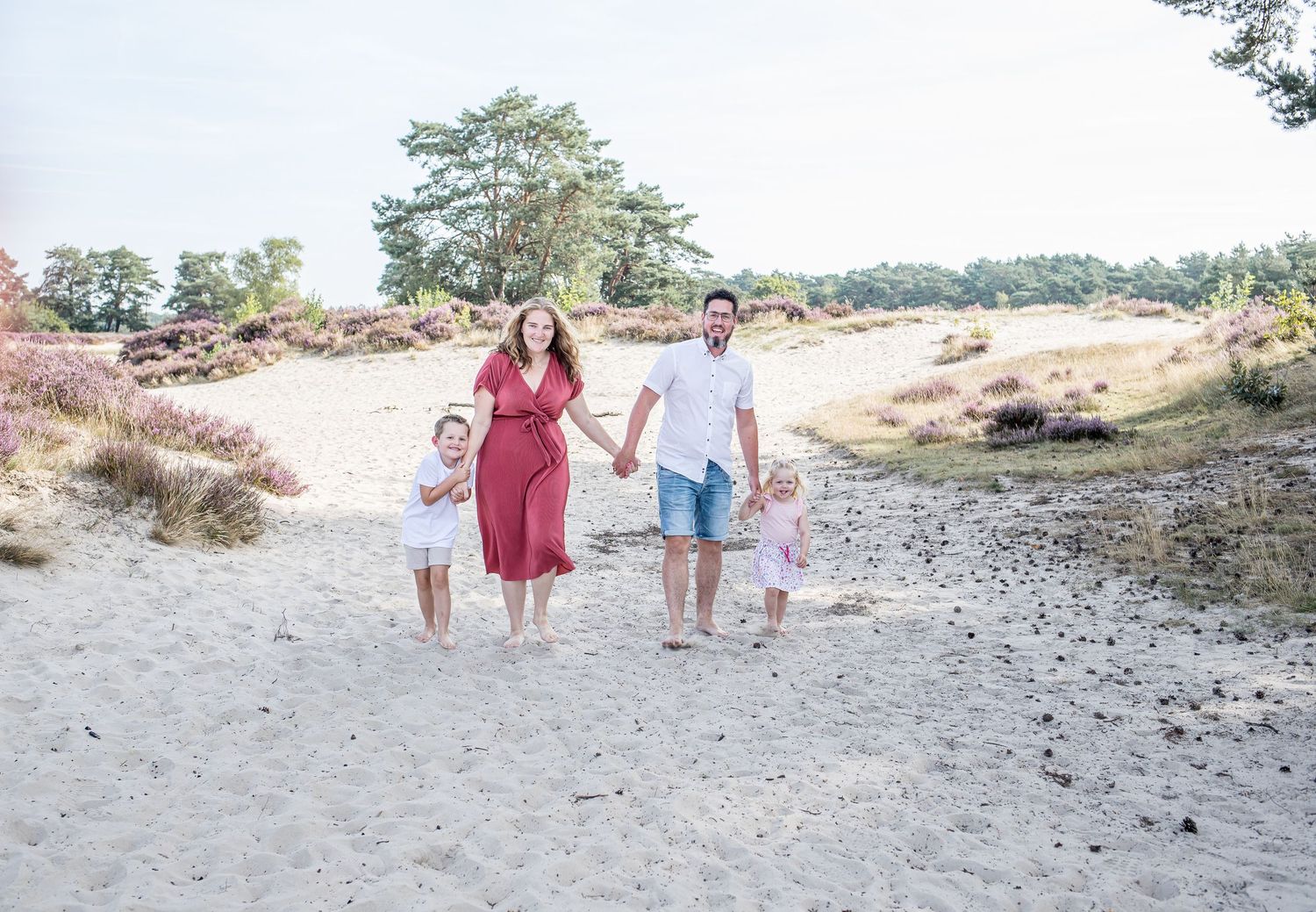Joyful family of four walking hand in hand on sandy path surrounded by heathland.