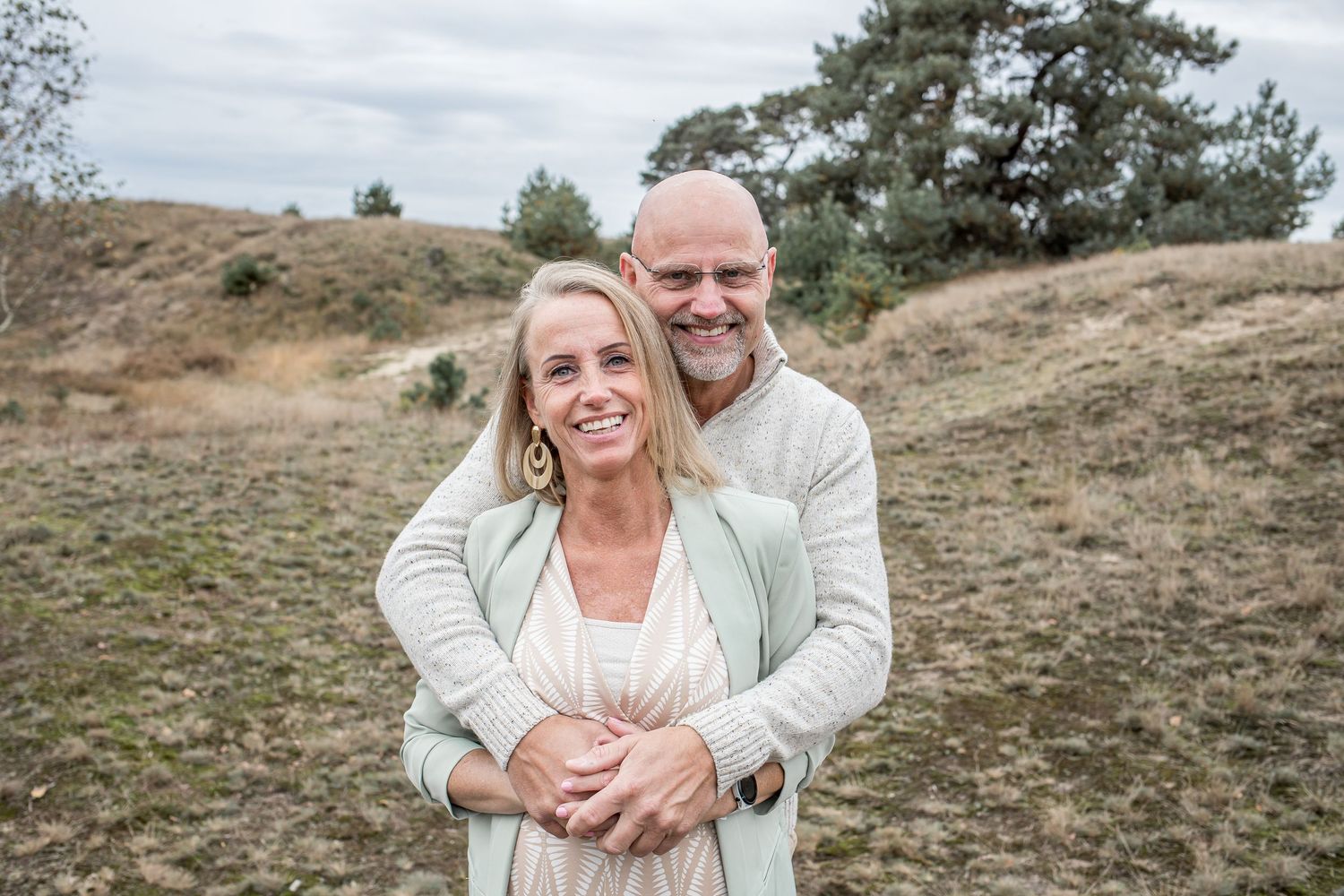 Smiling mature couple embracing outdoors on a hillside with pine trees behind them.
