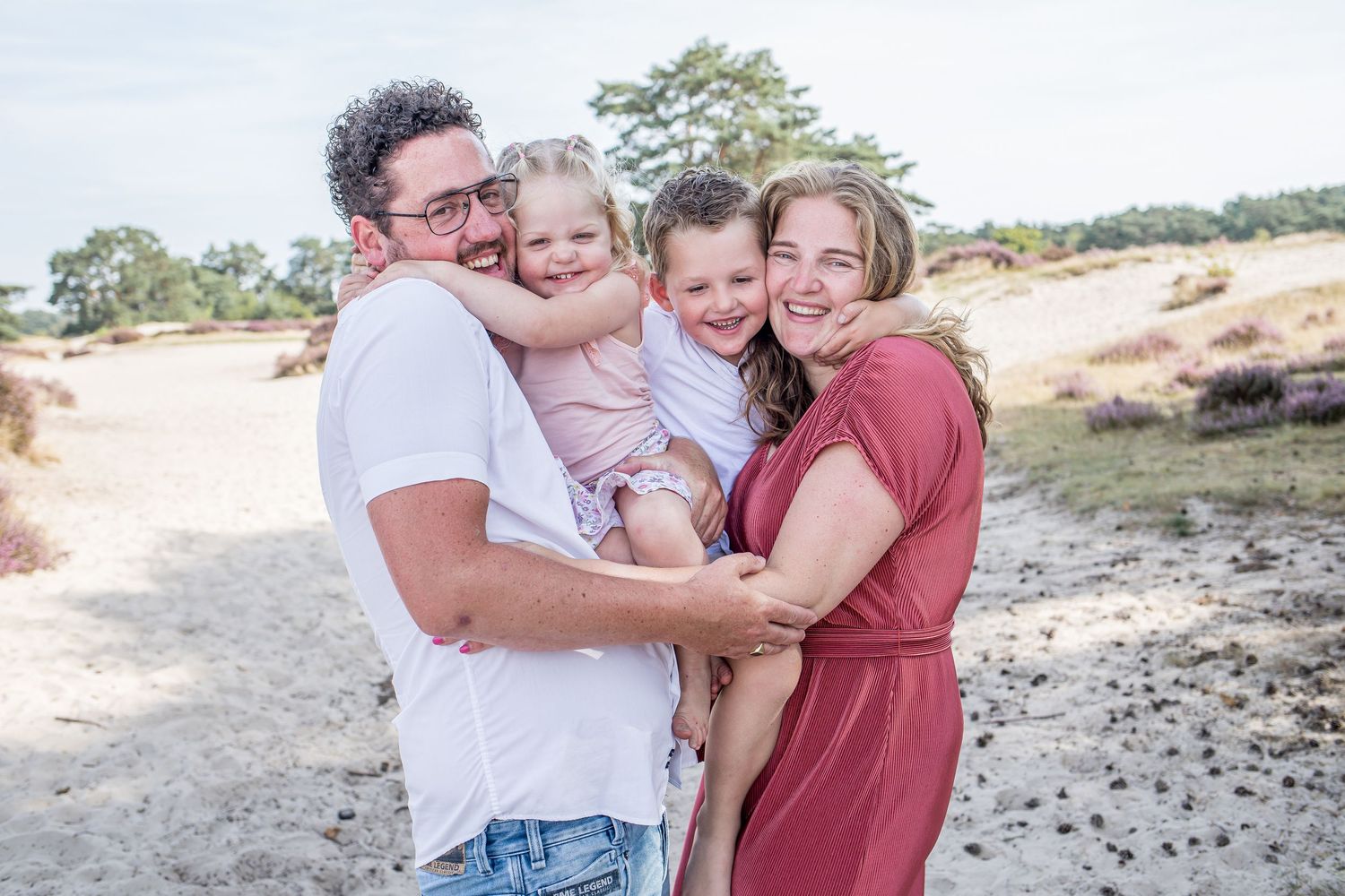 Happy family of four posing outdoors on sandy dunes, parents holding two young children.