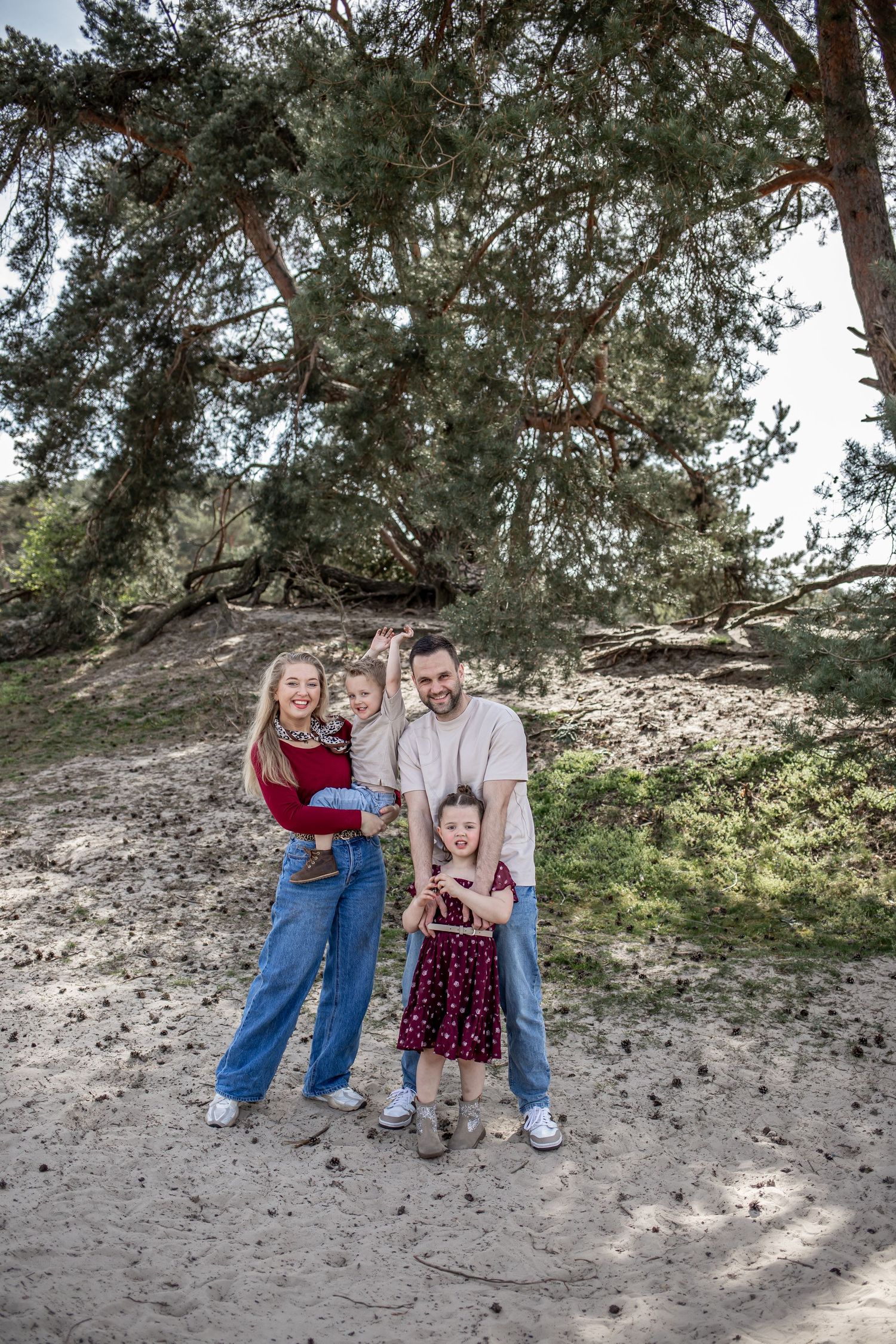 Happy family of four posing outdoors on sandy ground beneath large pine trees, parents holding young children.