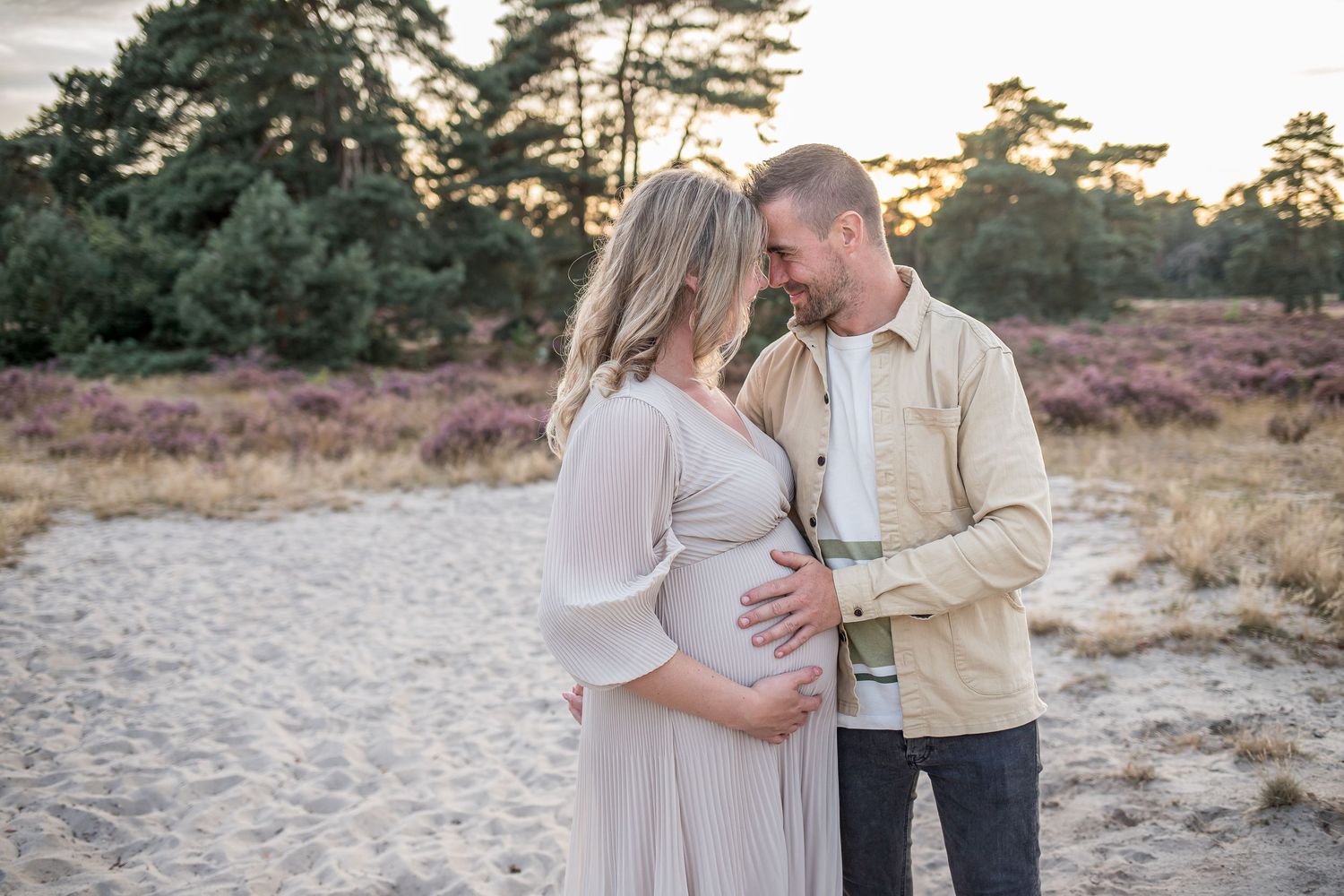 Expecting couple sharing tender moment on sandy heathland, man gently holding pregnant woman's belly at sunset.