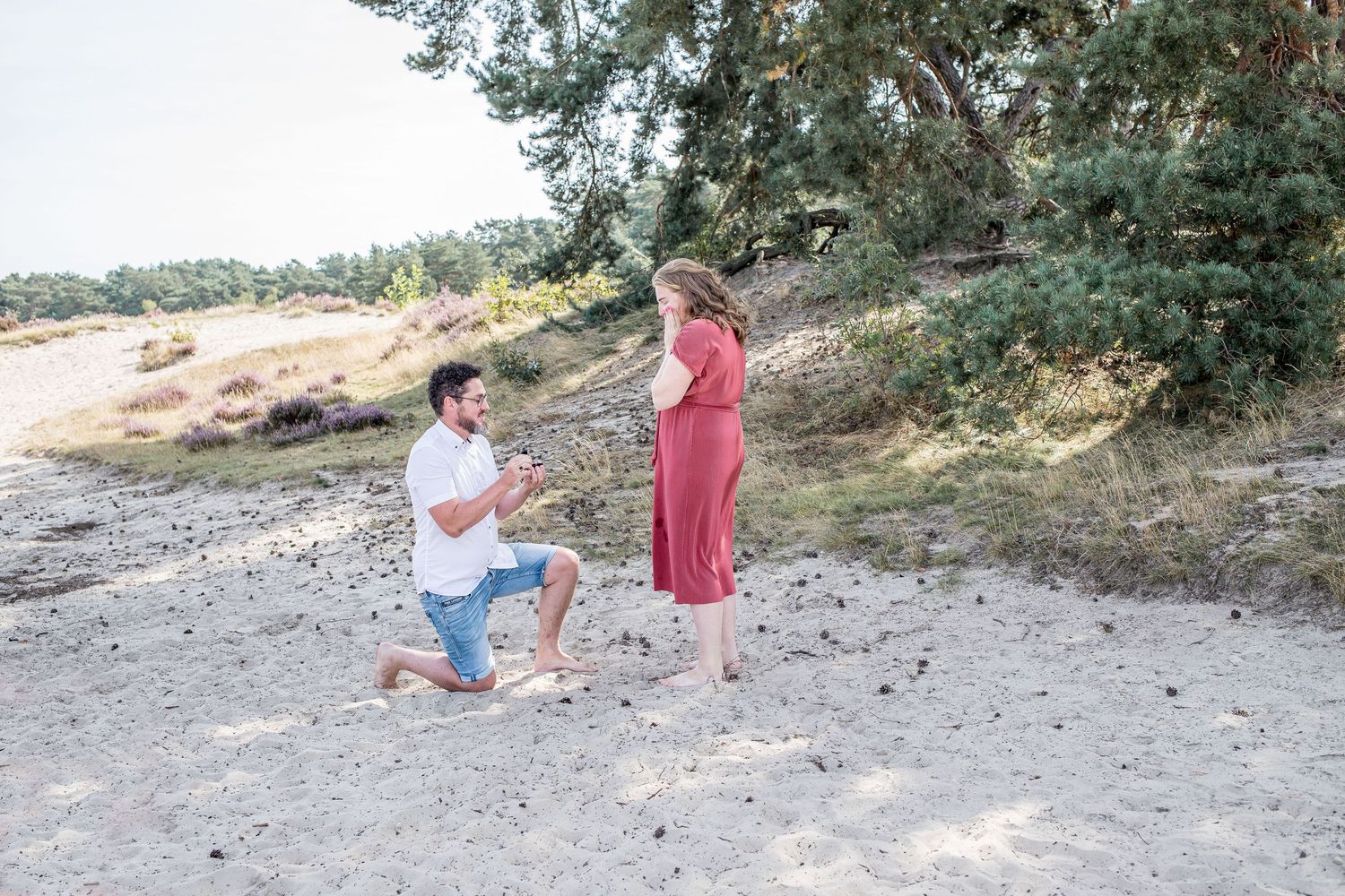 Man kneeling on sandy ground proposing to woman in red dress outdoors.