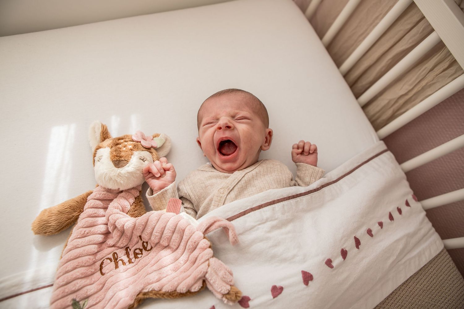 Newborn baby yawning in a white crib beside a personalized Chloe stuffed animal comforter.