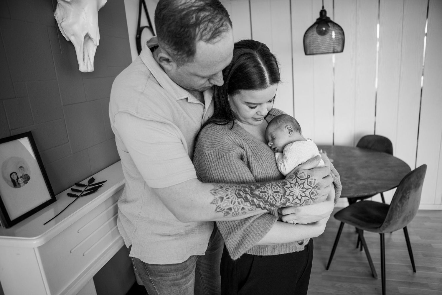 Black and white photo of new parents tenderly holding their swaddled newborn baby at home.