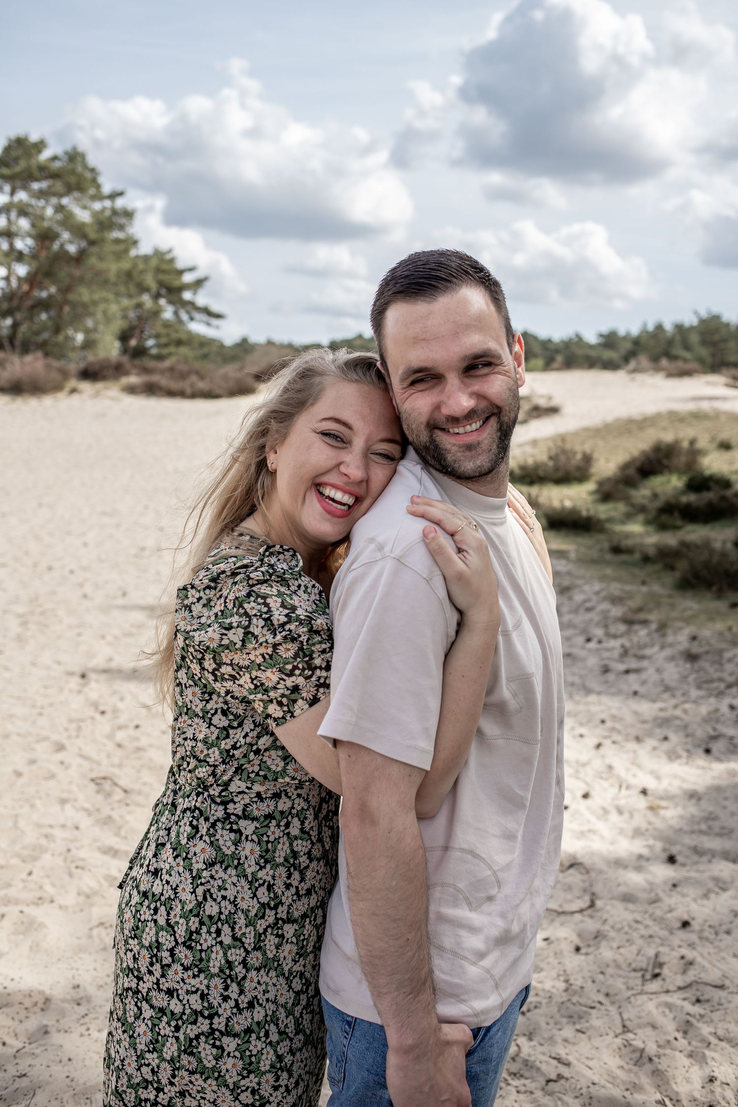 Happy couple embracing on sandy dunes, woman in floral dress leaning on smiling man in white tee.