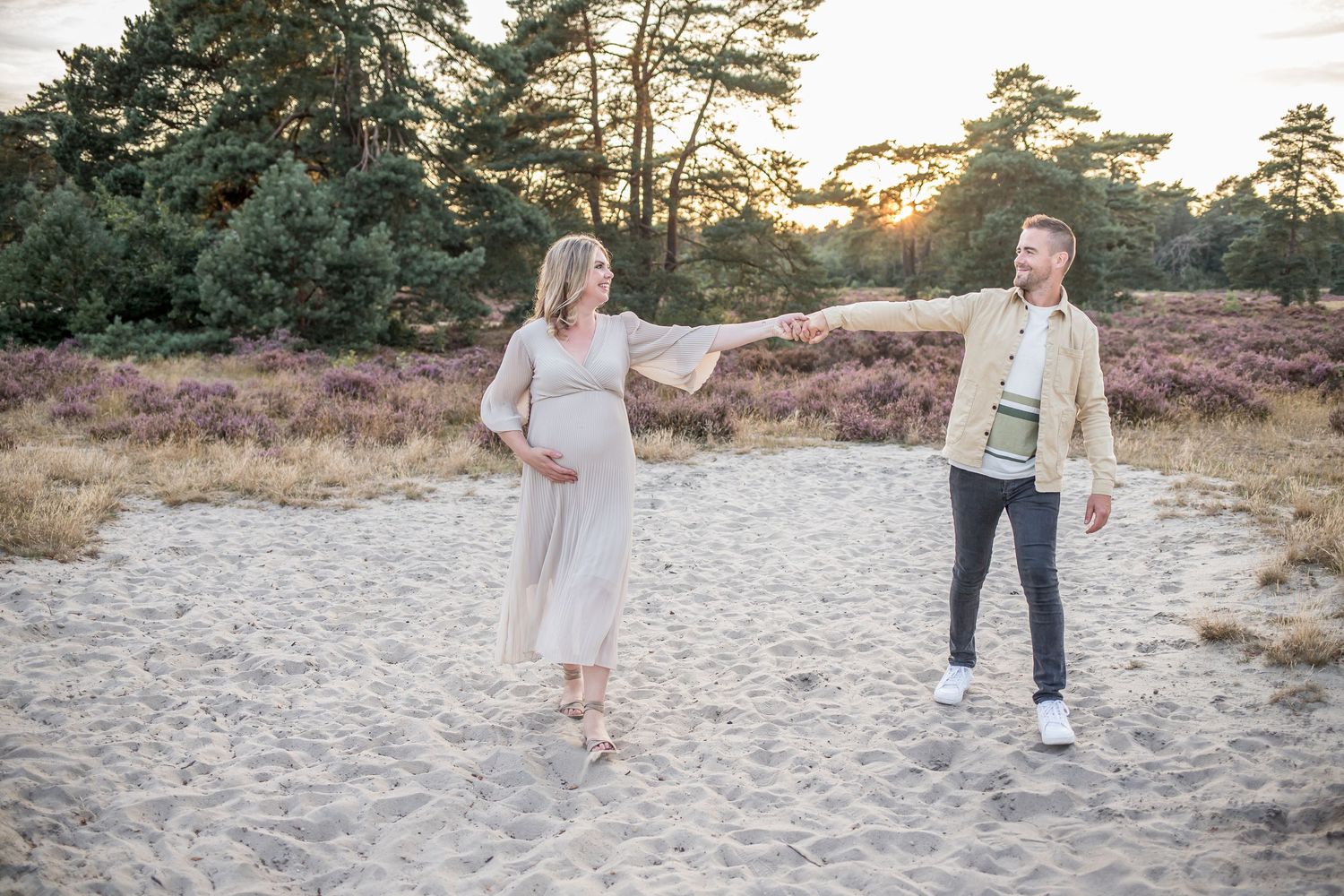 Pregnant woman and partner playfully reaching toward each other on a sandy path in heather fields.