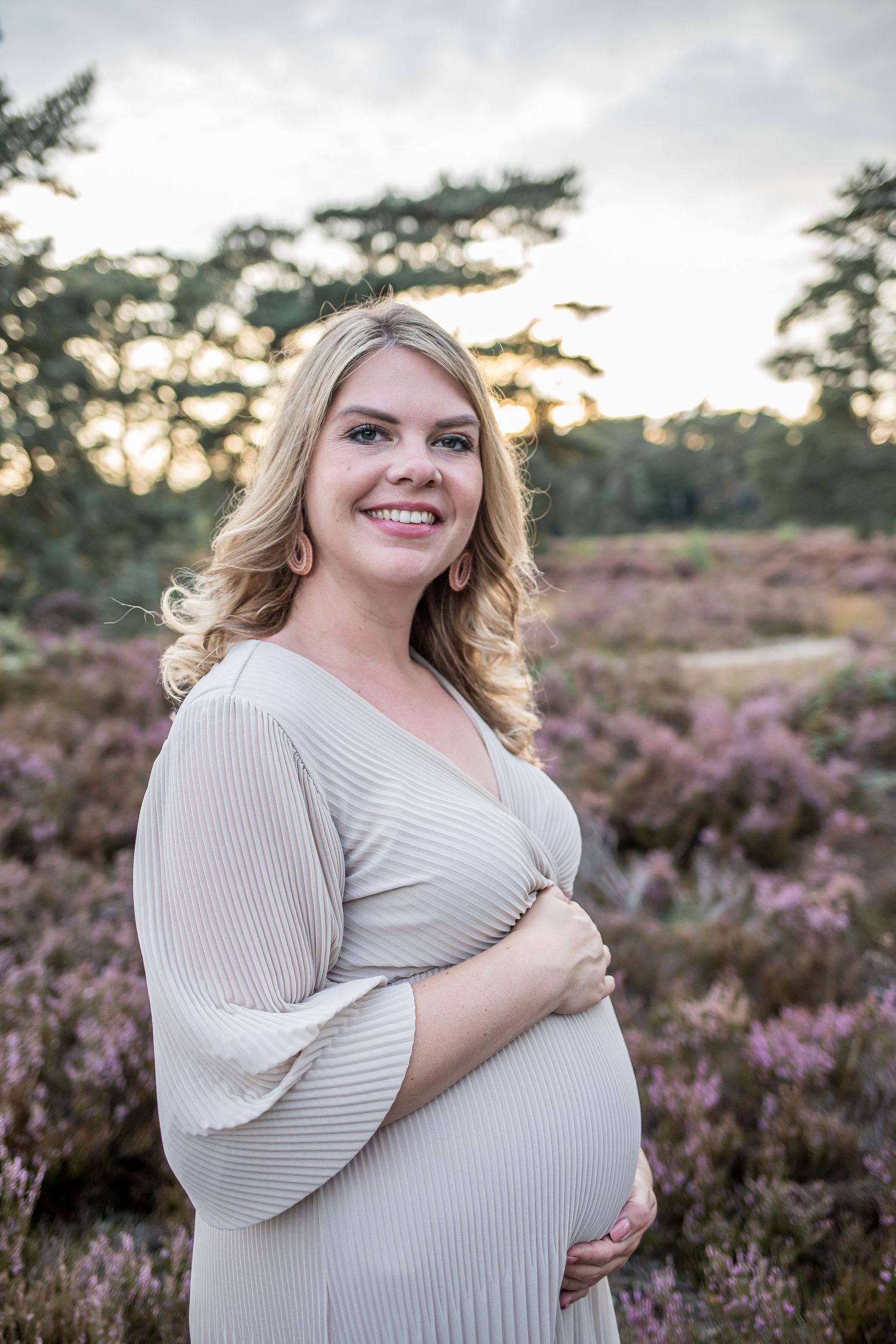 Happy pregnant woman in pleated cream dress smiling at camera in blooming purple heather landscape.