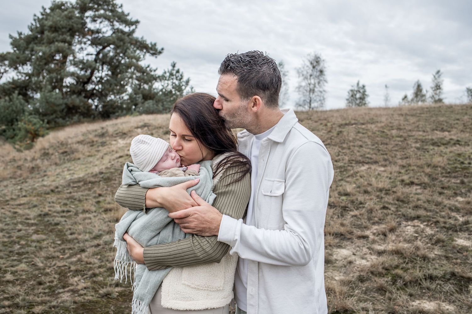 Loving couple tenderly holding newborn baby wrapped in blanket on hillside outdoors.