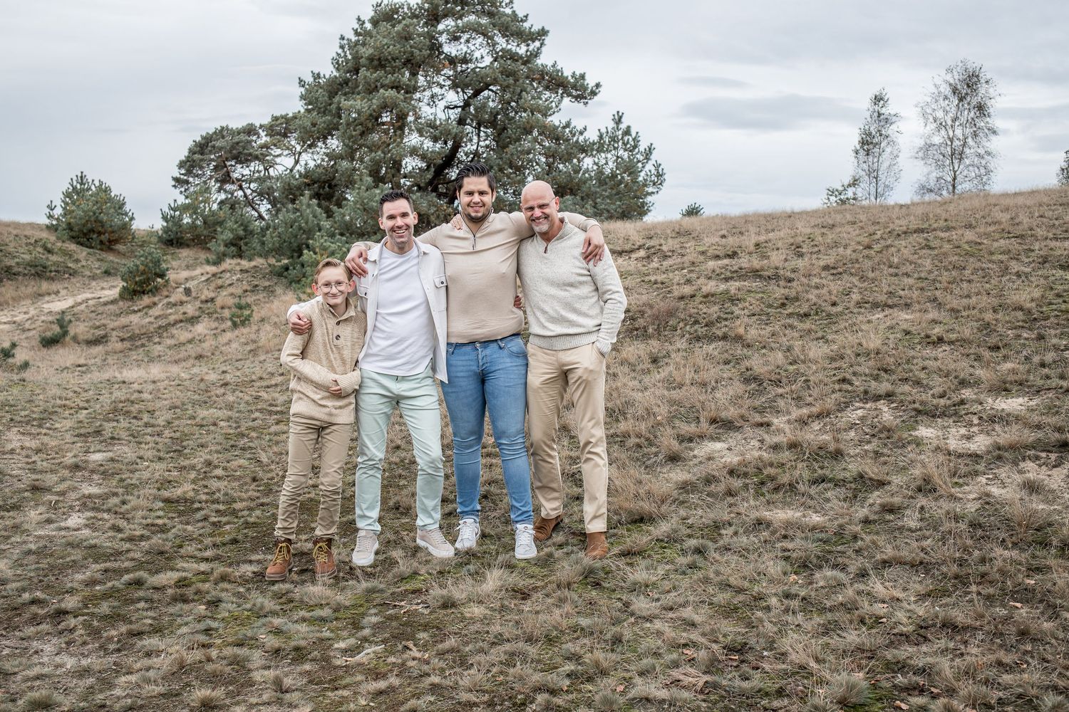 Four men stand together smiling on a grassy hillside with pine trees behind them.