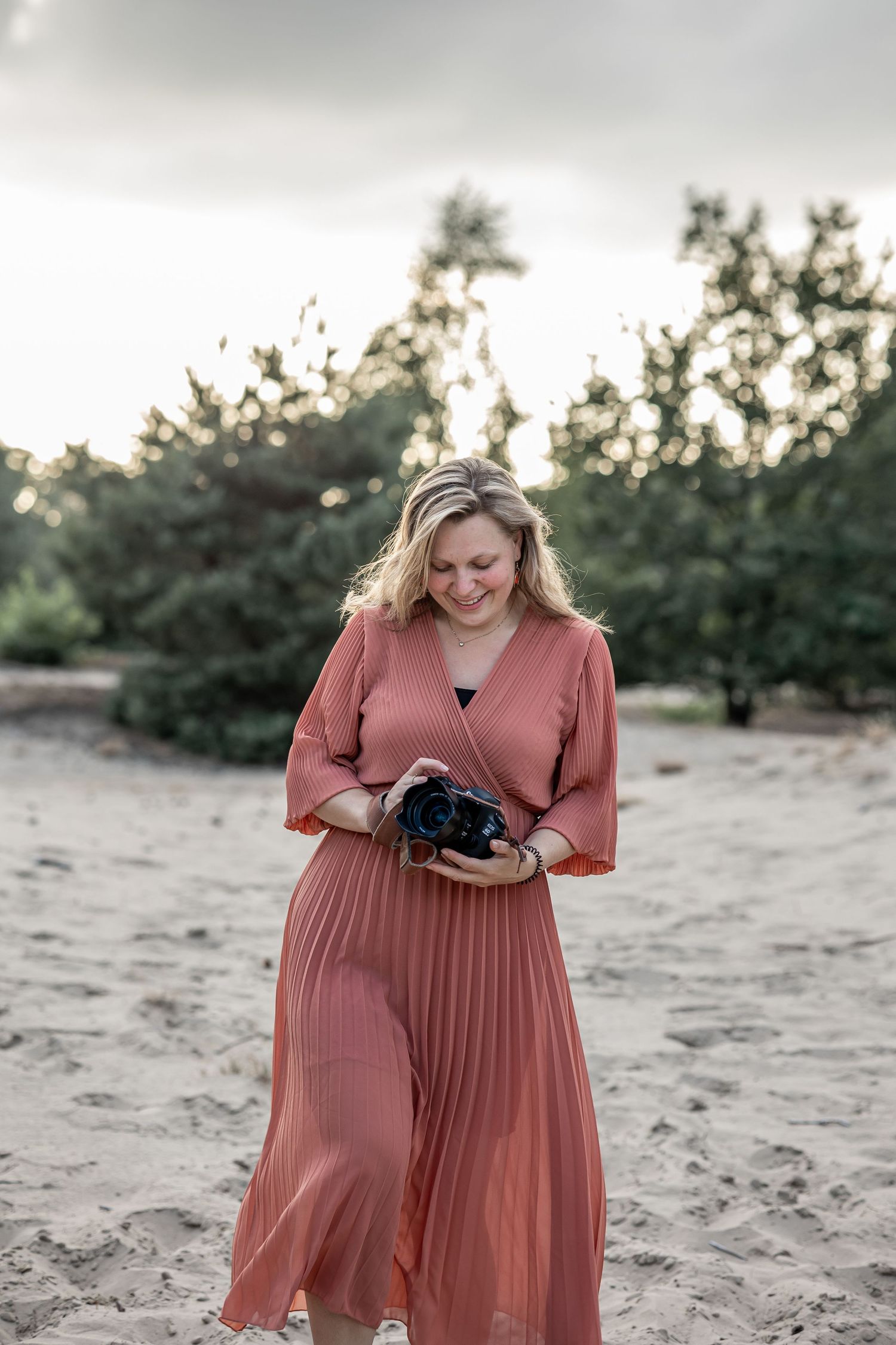 Blonde woman in rust pleated dress reviewing photos on camera outdoors.