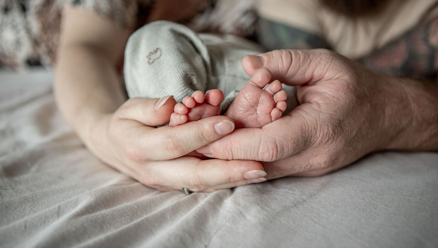 Close-up of parents gently cradling tiny newborn baby feet in their hands on a soft white blanket.