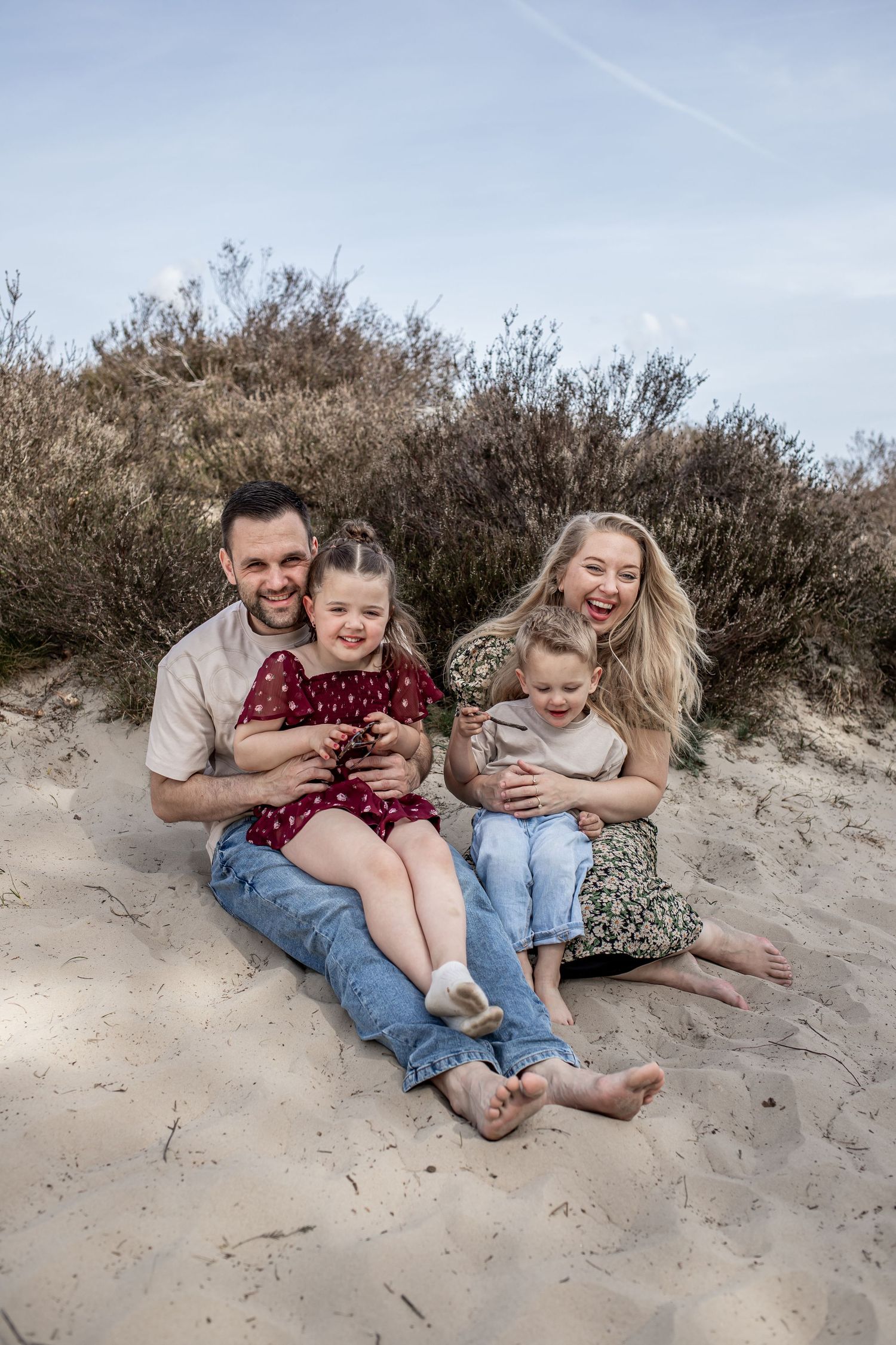 Happy family of four sitting on sandy dunes, parents hugging two young children, smiling outdoors with shrubs in background.