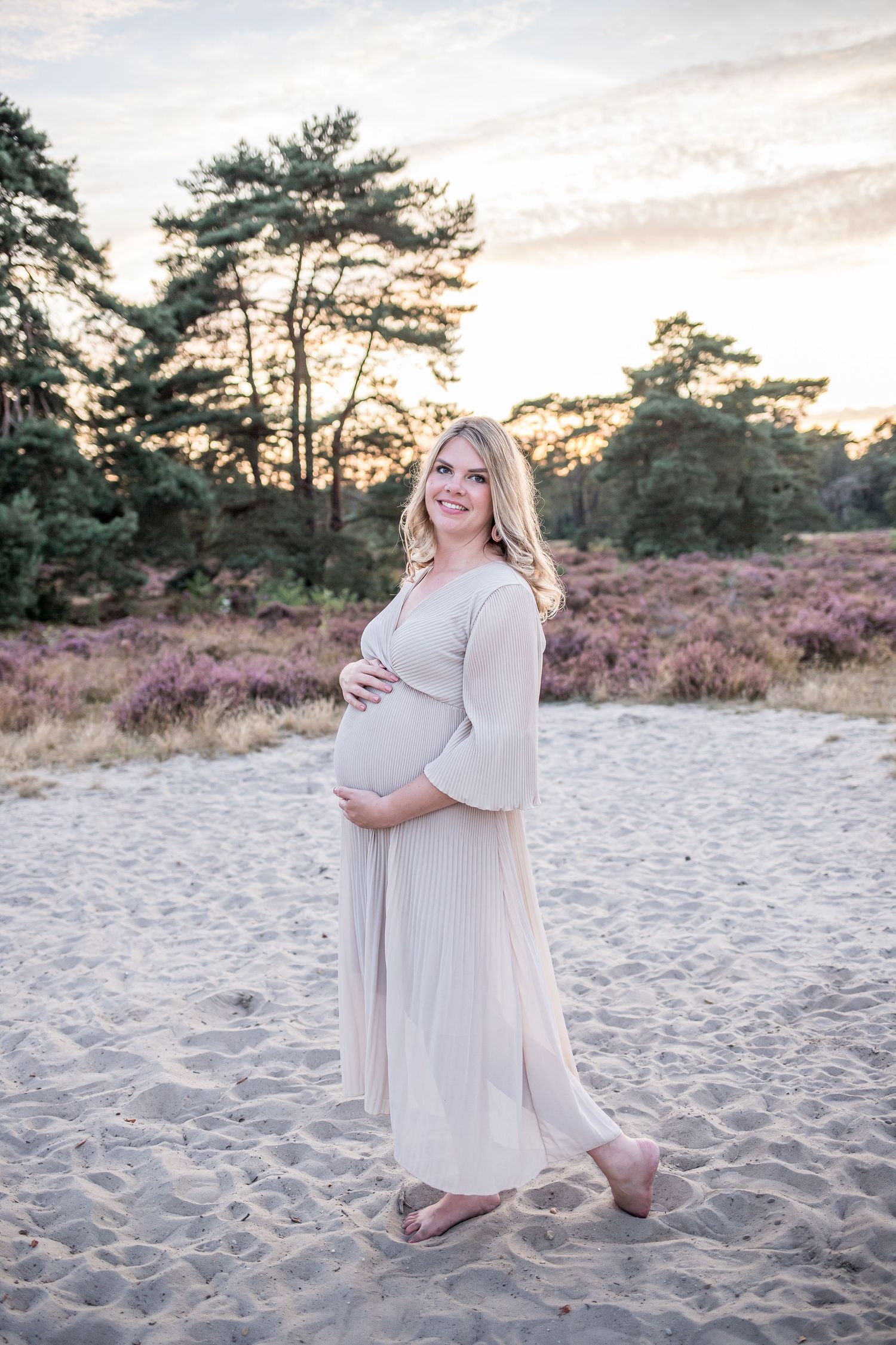 Smiling pregnant woman in elegant cream maternity gown standing barefoot on sandy heathland at dusk.