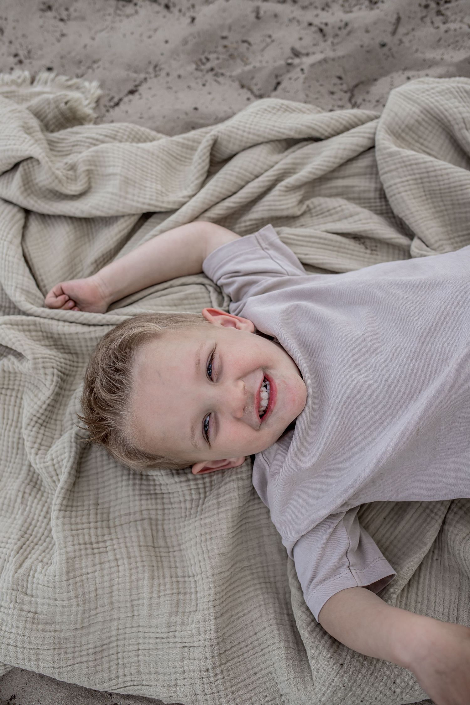 Smiling young child lying on a beige textured blanket on sandy ground, looking up happily.