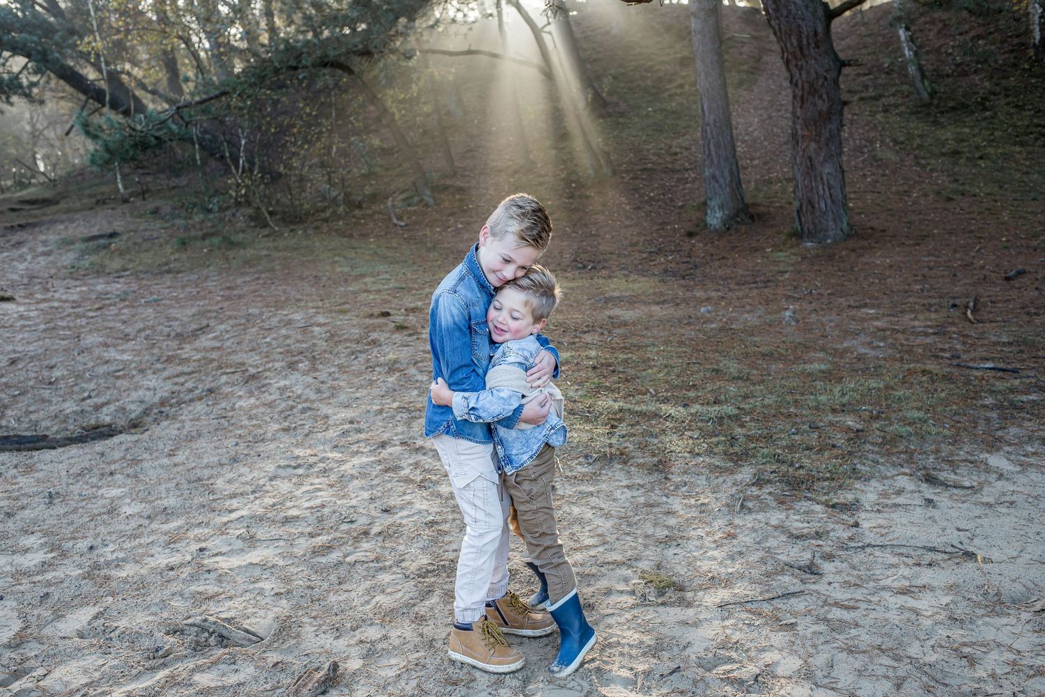 Two young children hugging on sandy forest path with sunbeams shining through trees.