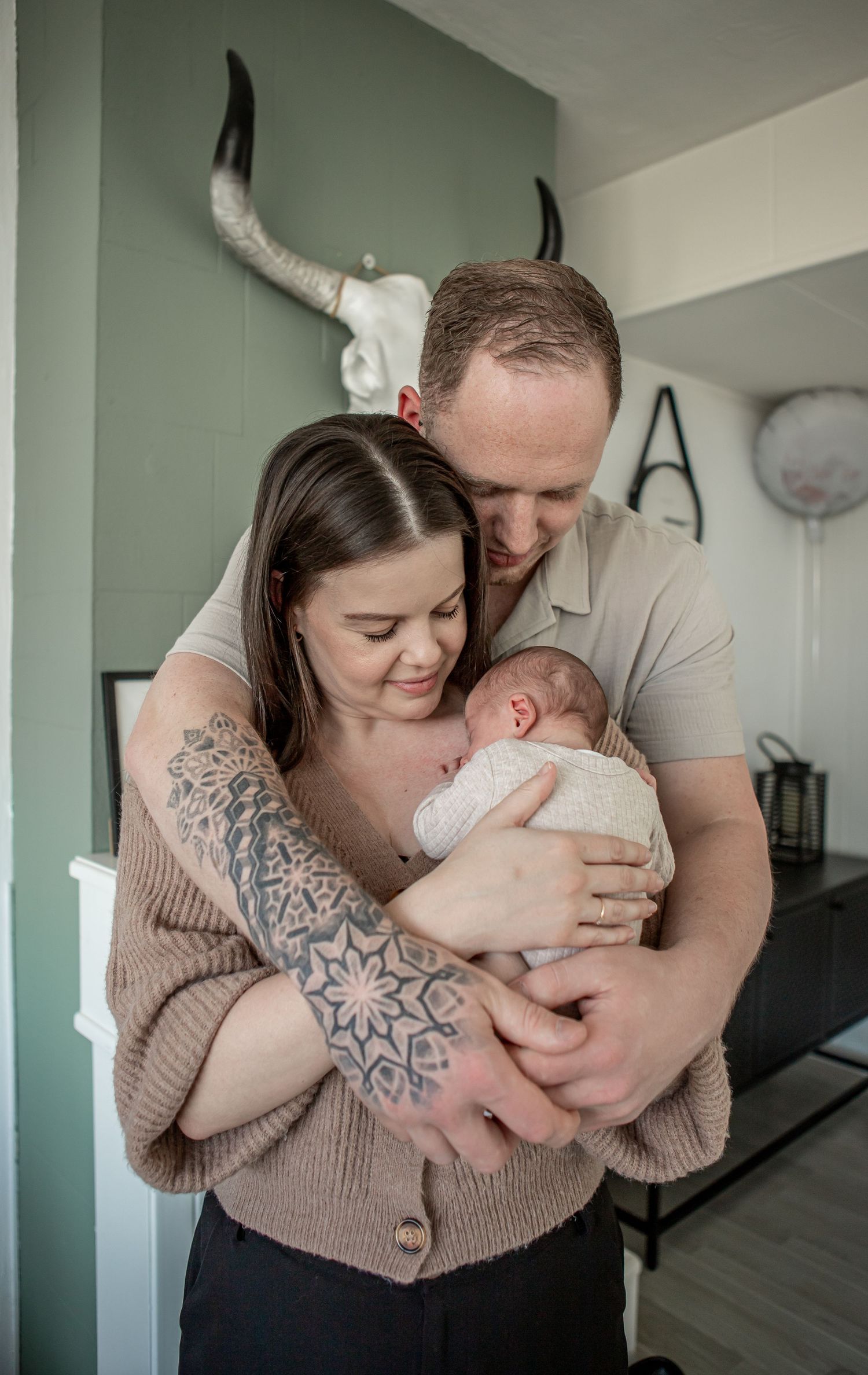 Tattooed mother and father holding their swaddled newborn baby together in a green-walled home.