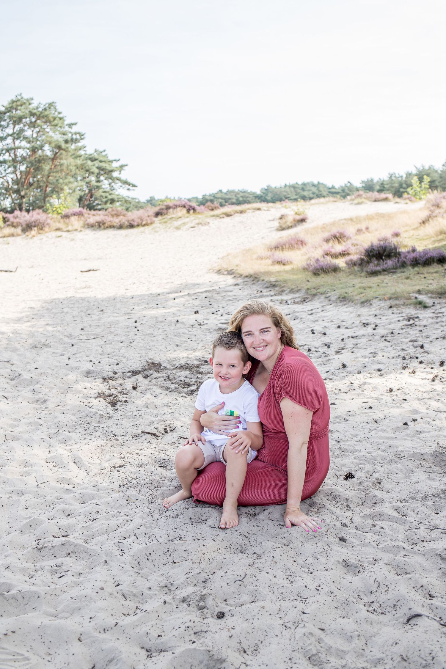 A smiling mother in a red dress sits on sandy ground cuddling her young toddler son outdoors.