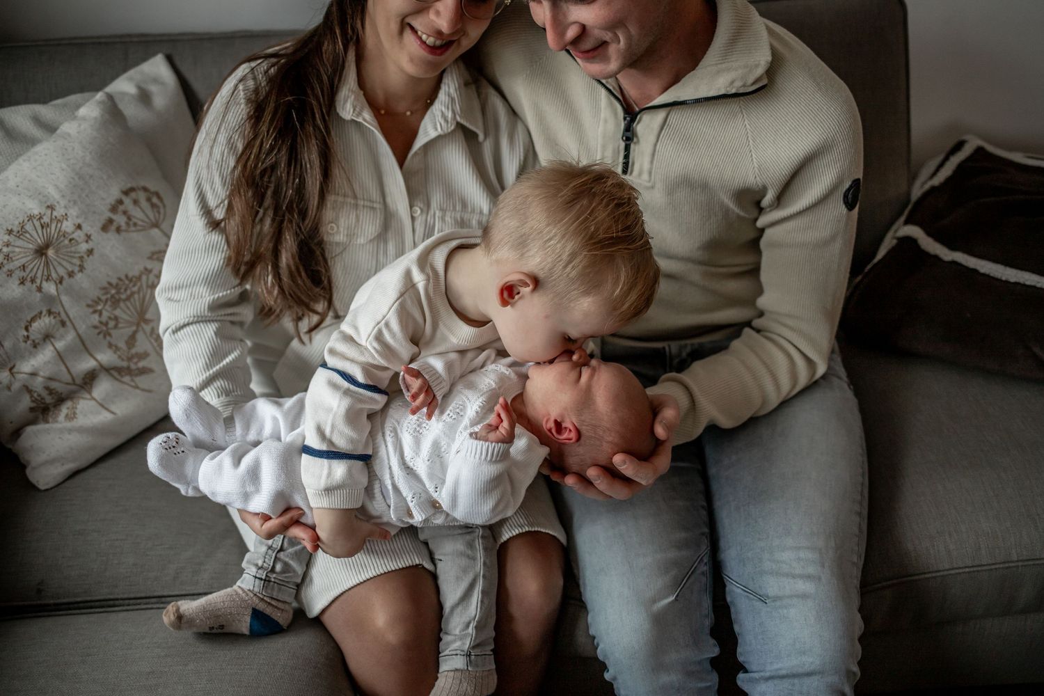 Parents sitting on a couch smiling as their toddler gently holds a newborn baby sibling.