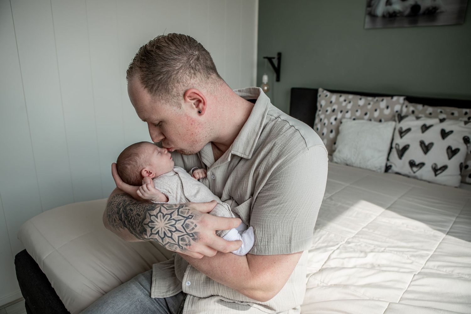 Tattooed father lovingly kissing his newborn baby while cradling them in a bedroom.