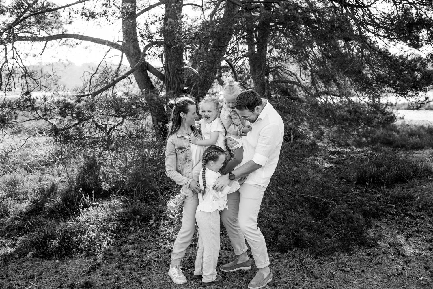 Black and white family photo with parents and children playing together under pine trees outdoors.