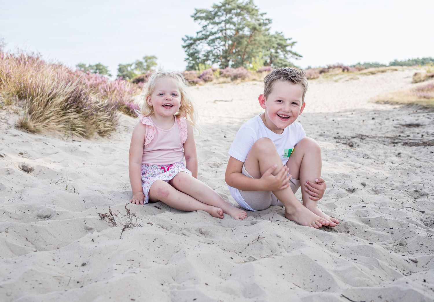 A smiling young girl and boy sit together on sandy ground with purple heather and trees in the background.