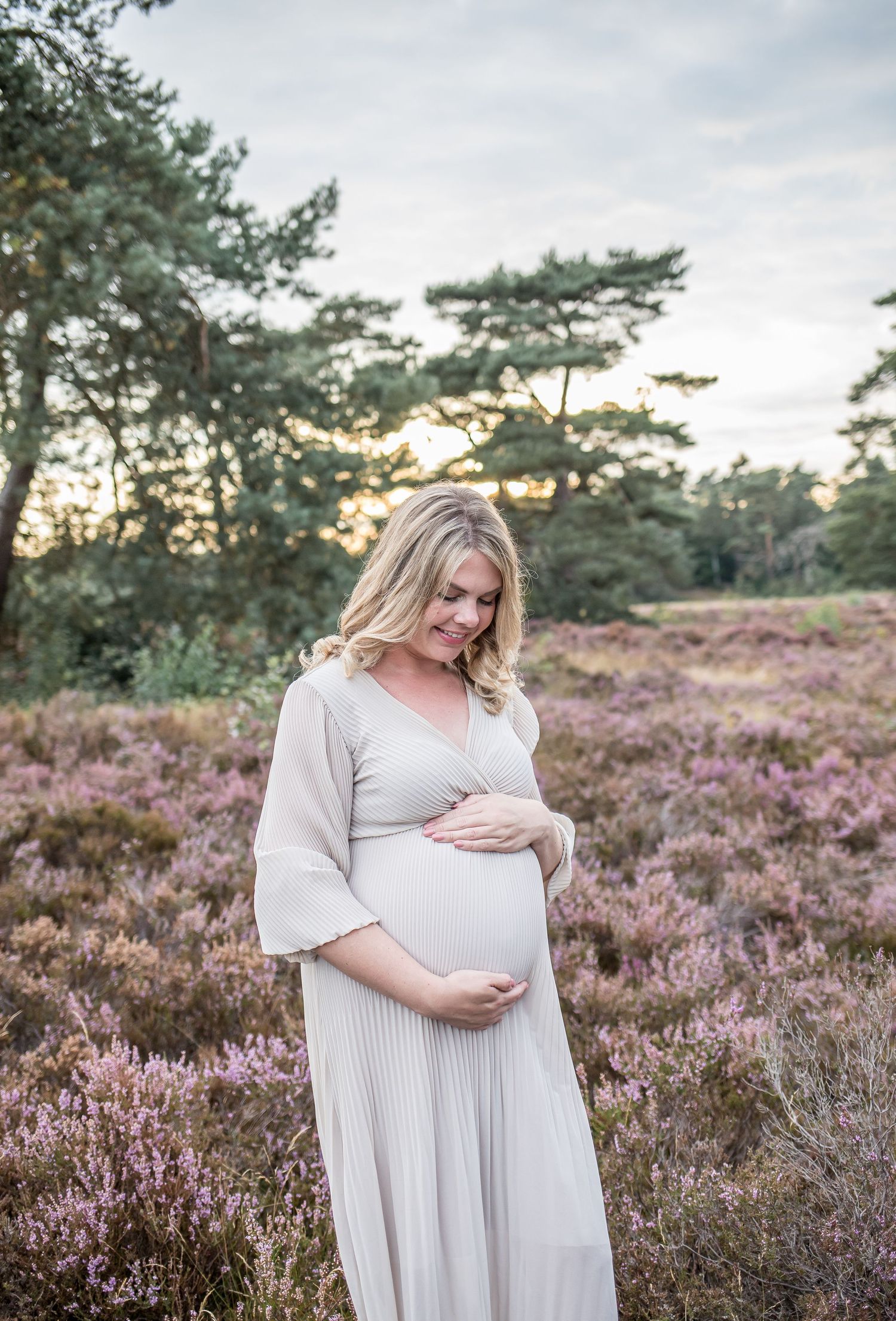 Pregnant blonde woman in white dress cradling baby bump among purple heather blooms at sunset.