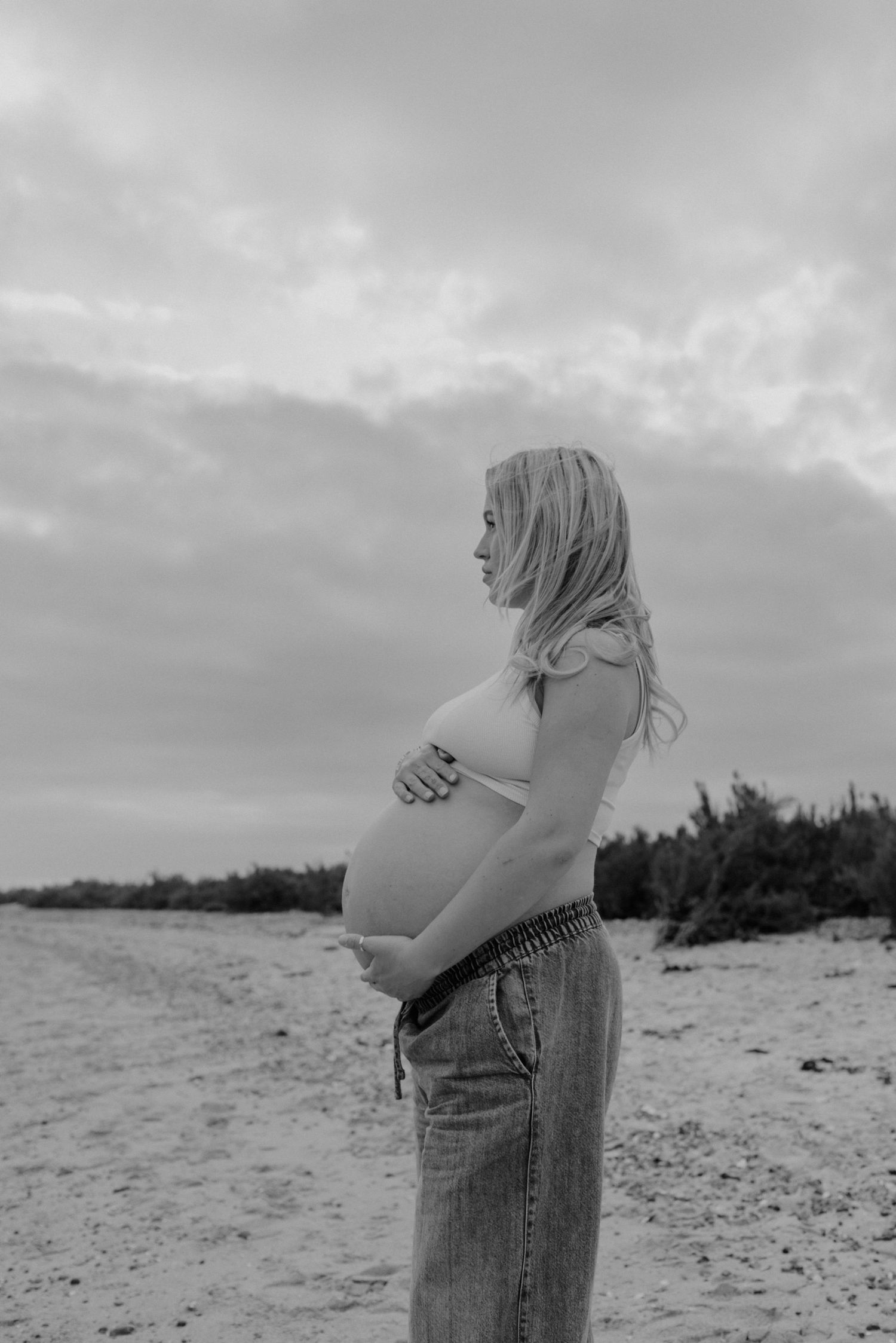 A black and white beach maternity photo capturing a silhouette against a cloudy sky.