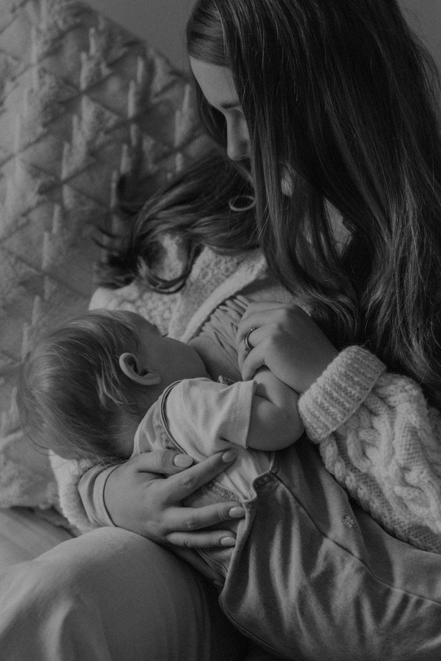 A tender black and white photograph of a mother nursing her baby in a cozy sweater against a quilted backdrop.