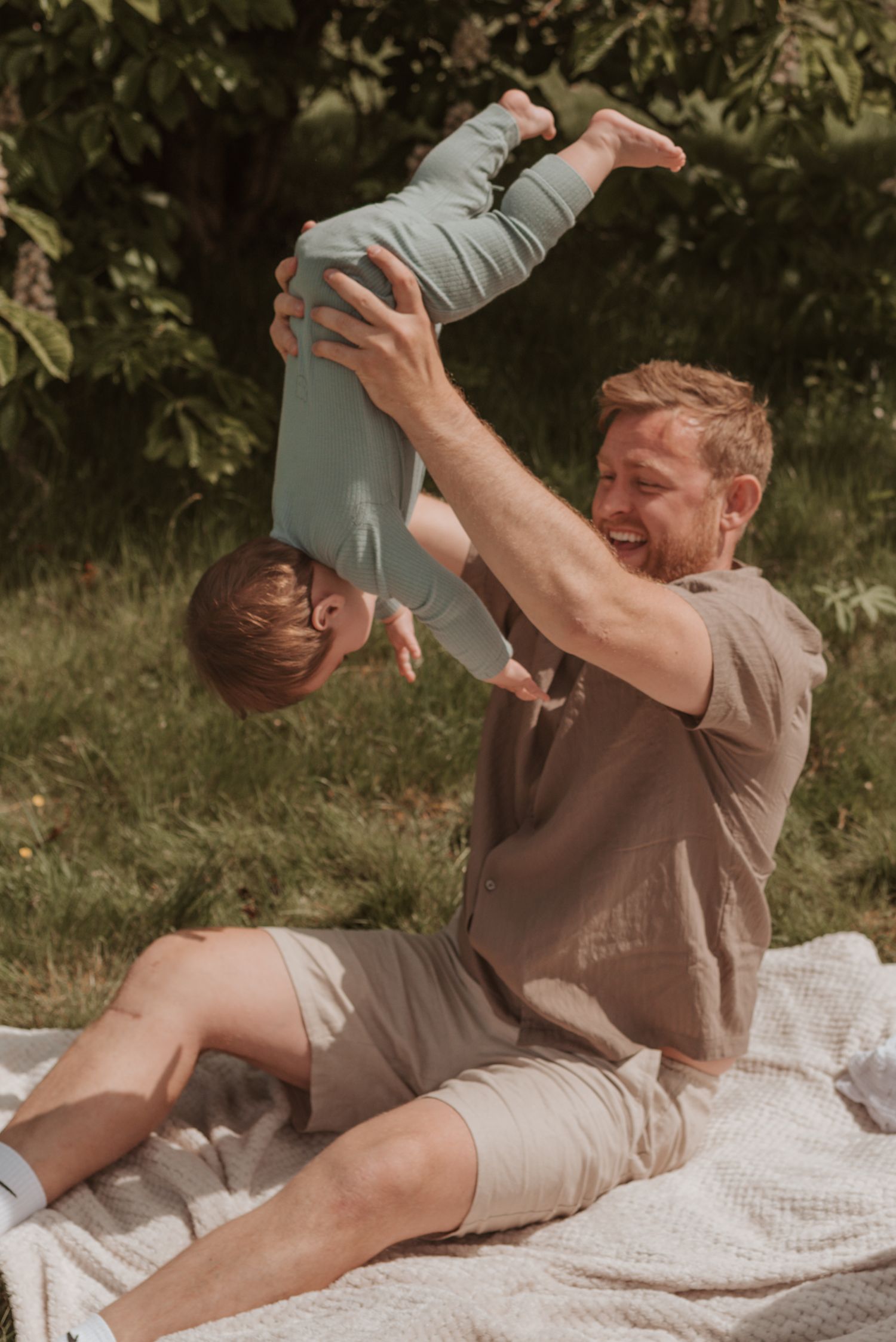 Playful moment captured outdoors on blanket with joyful airplane-style lifting.