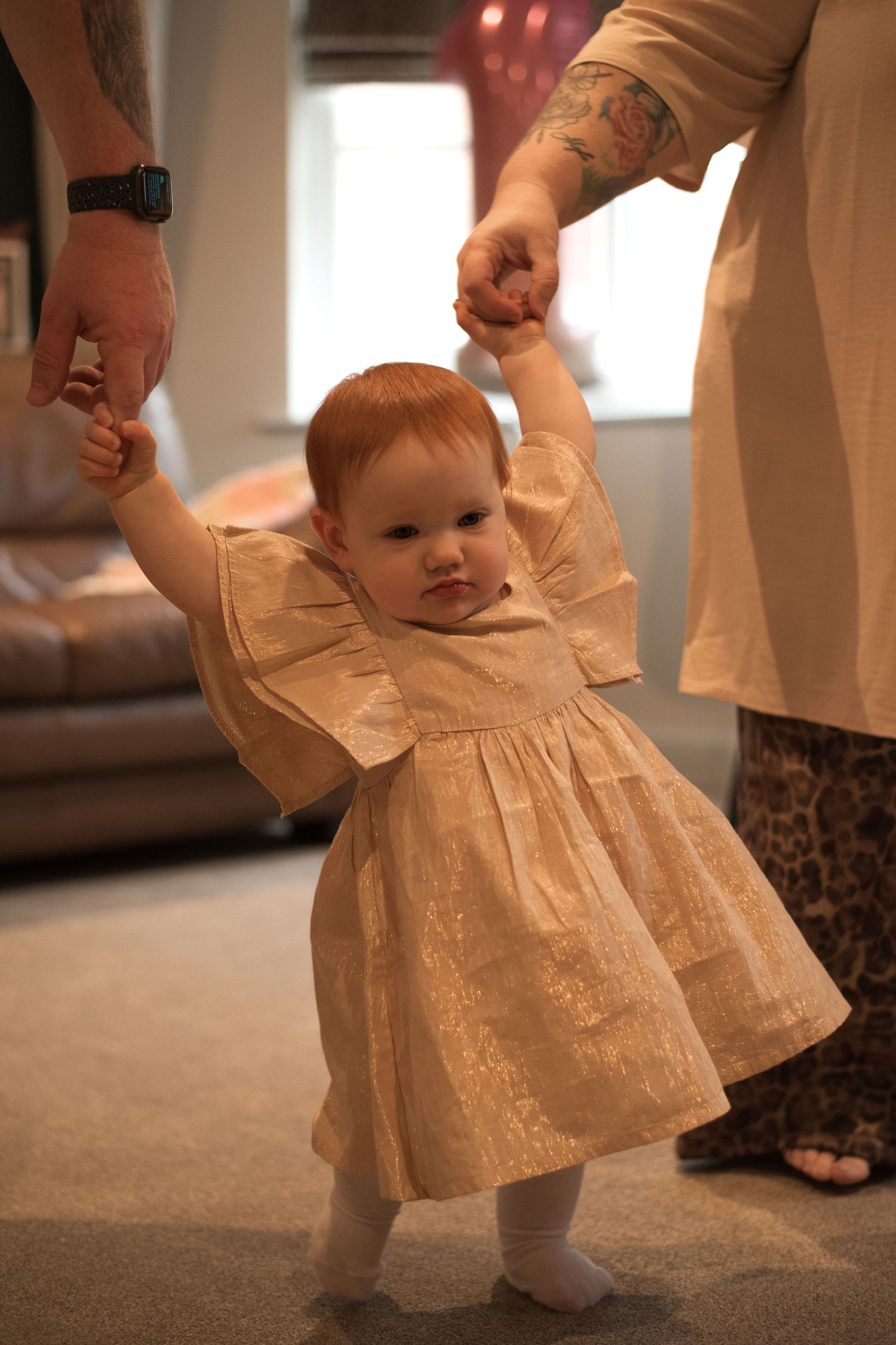 Small child in a cream-colored dress learning to walk while holding hands with adults.